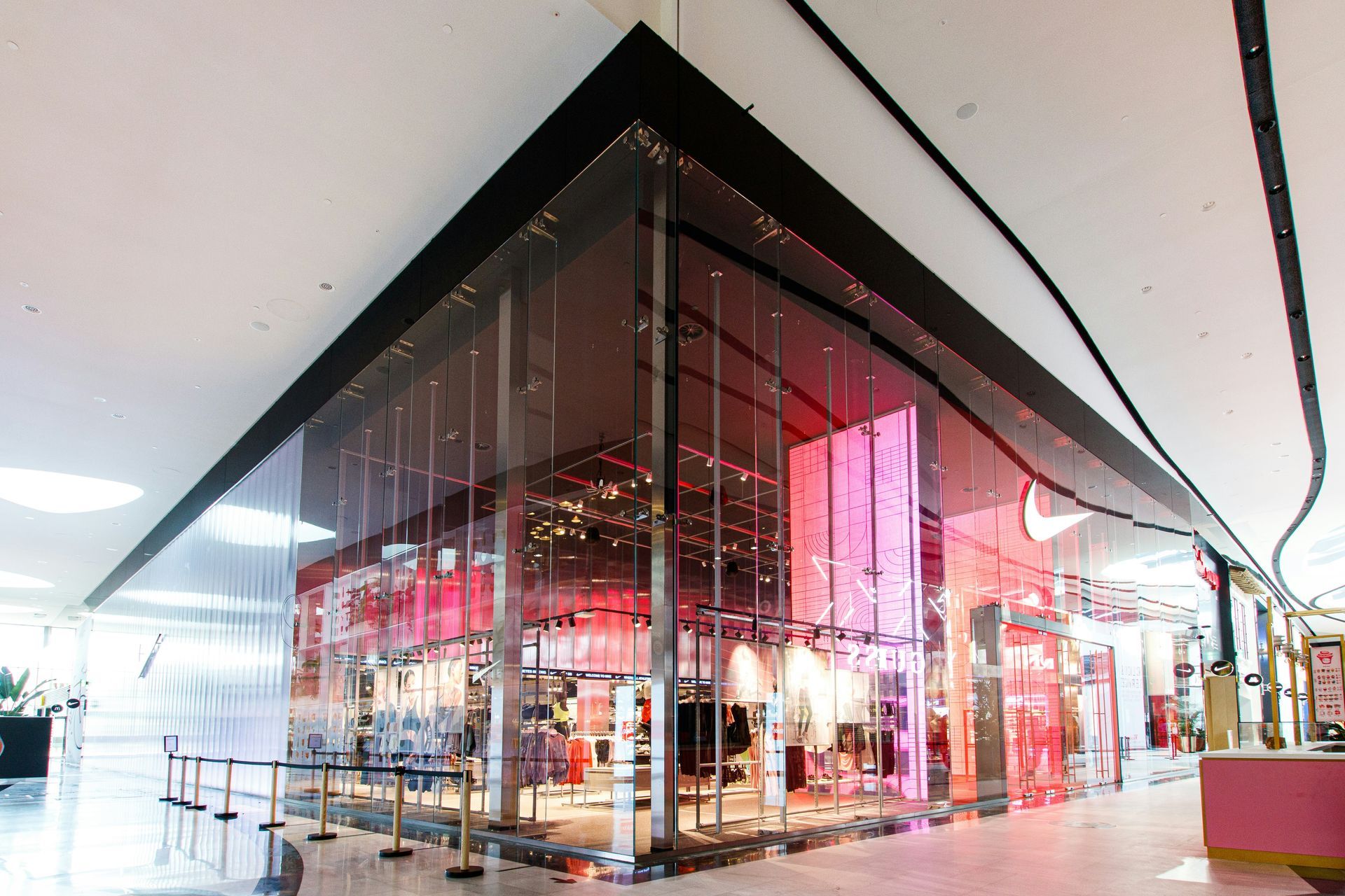 Nike store exterior, transparent glass walls, red and white interior, corner retail space, shopping mall setting.