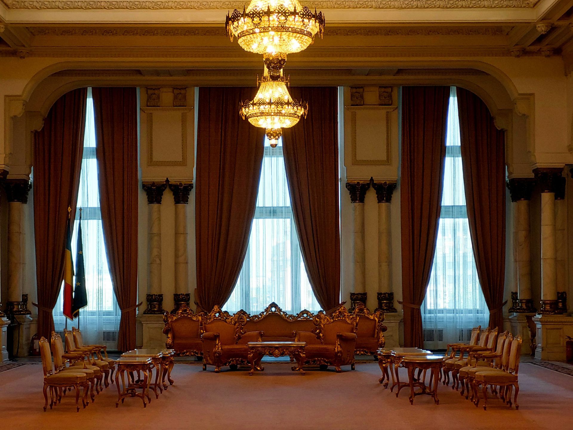 Formal room with ornate furniture, gold chandeliers, tall windows with brown drapes, and pink carpet.