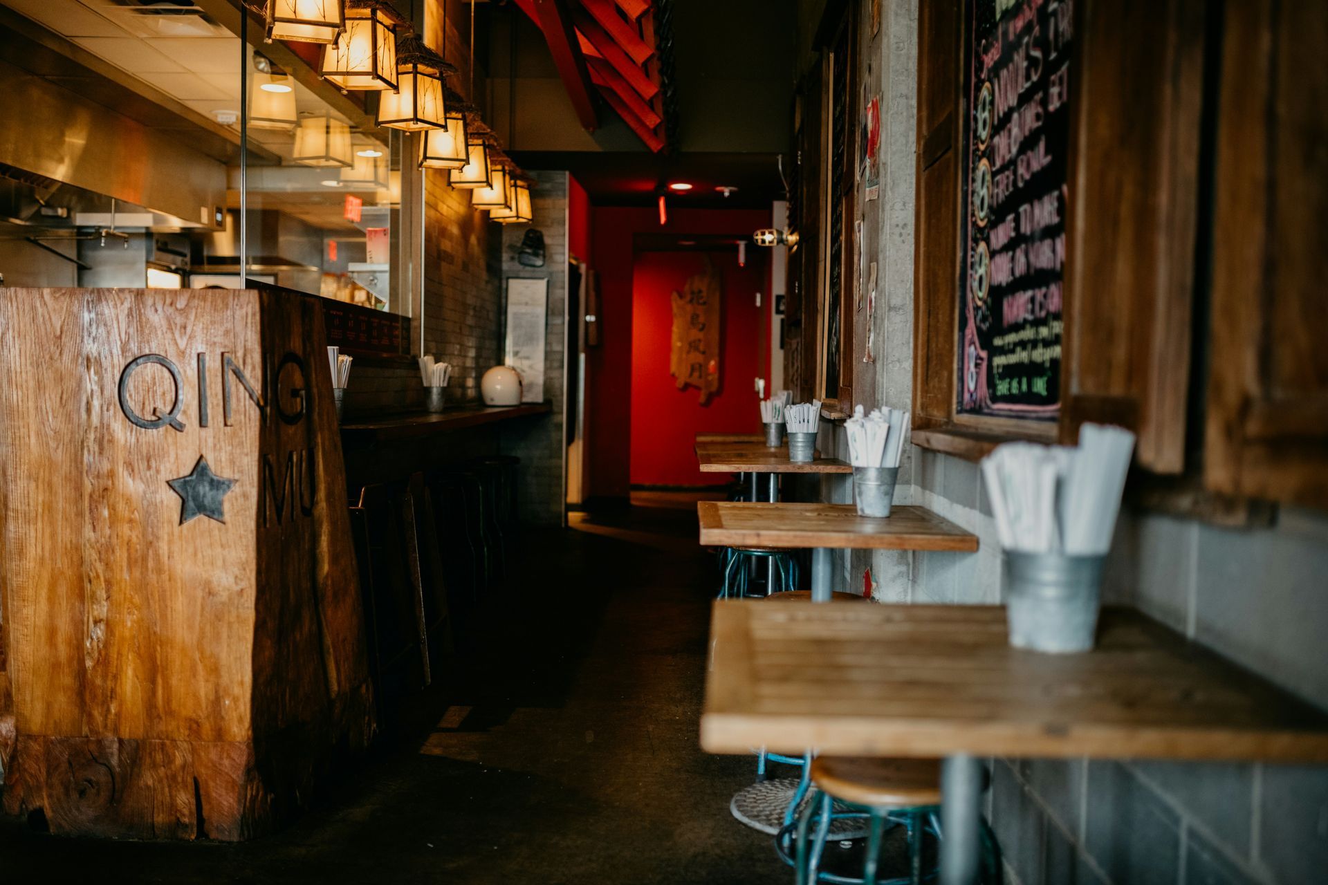Restaurant interior: wooden counter, tables with metal containers holding napkins, red doorway, overhead lighting.