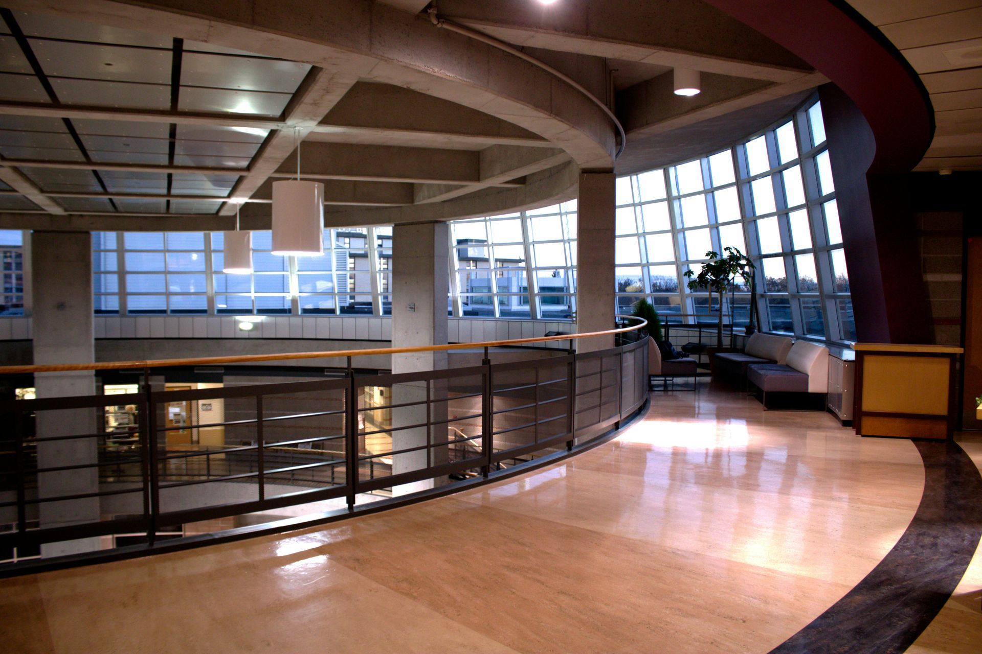 Interior view of a building's upper level. Curved hallway with large windows and a railing overlooks another floor.