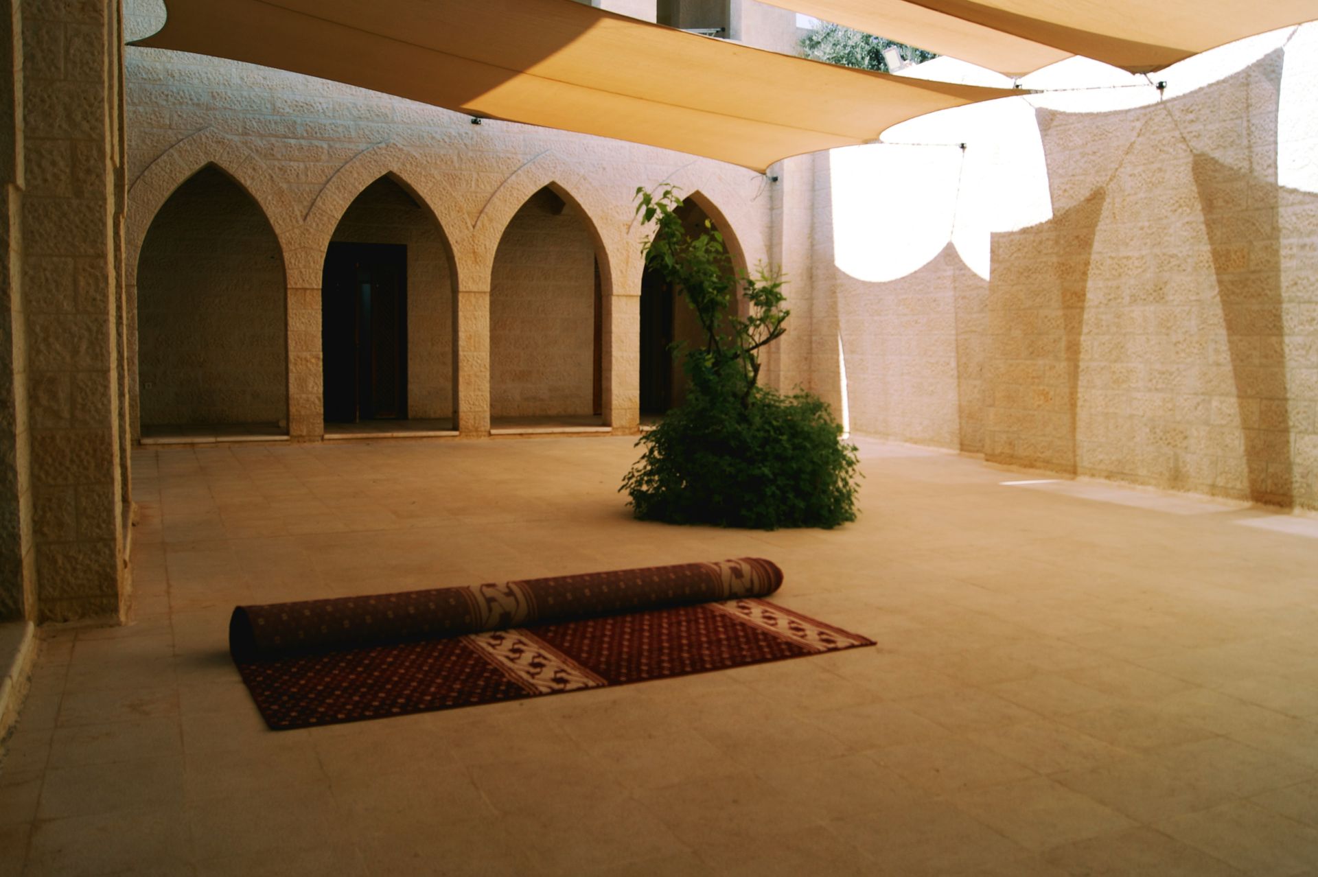 Courtyard with arches, beige walls, sunshade, rolled rug, and green plant.