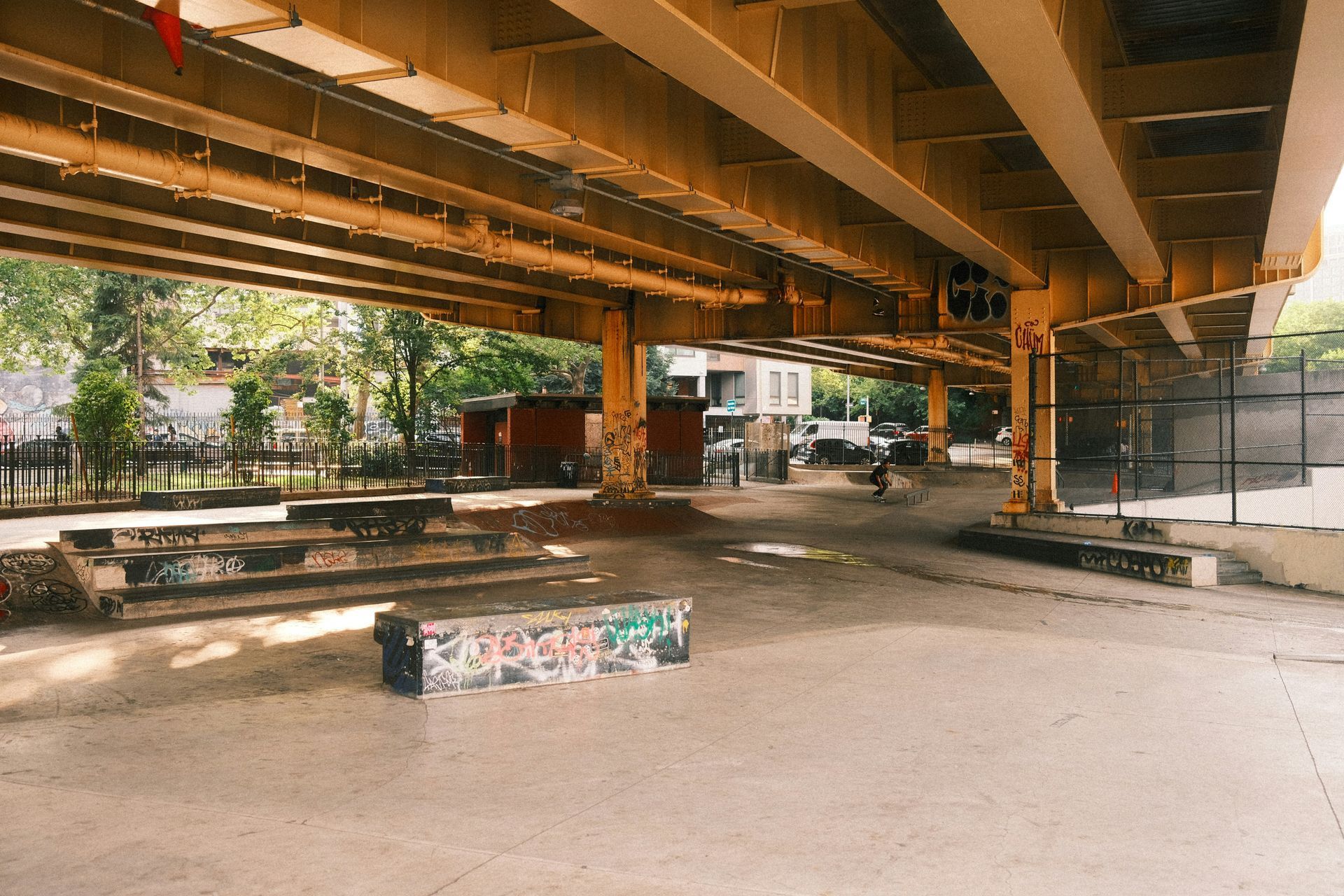 Skate park under a bridge with concrete ramps, graffiti-covered benches, and a fence; day.