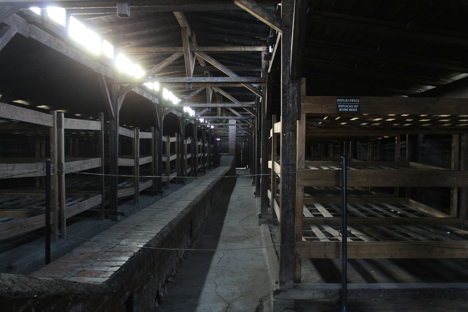 Interior of a wooden barrack with multiple-tiered bunk beds, long hallway, and dim lighting.