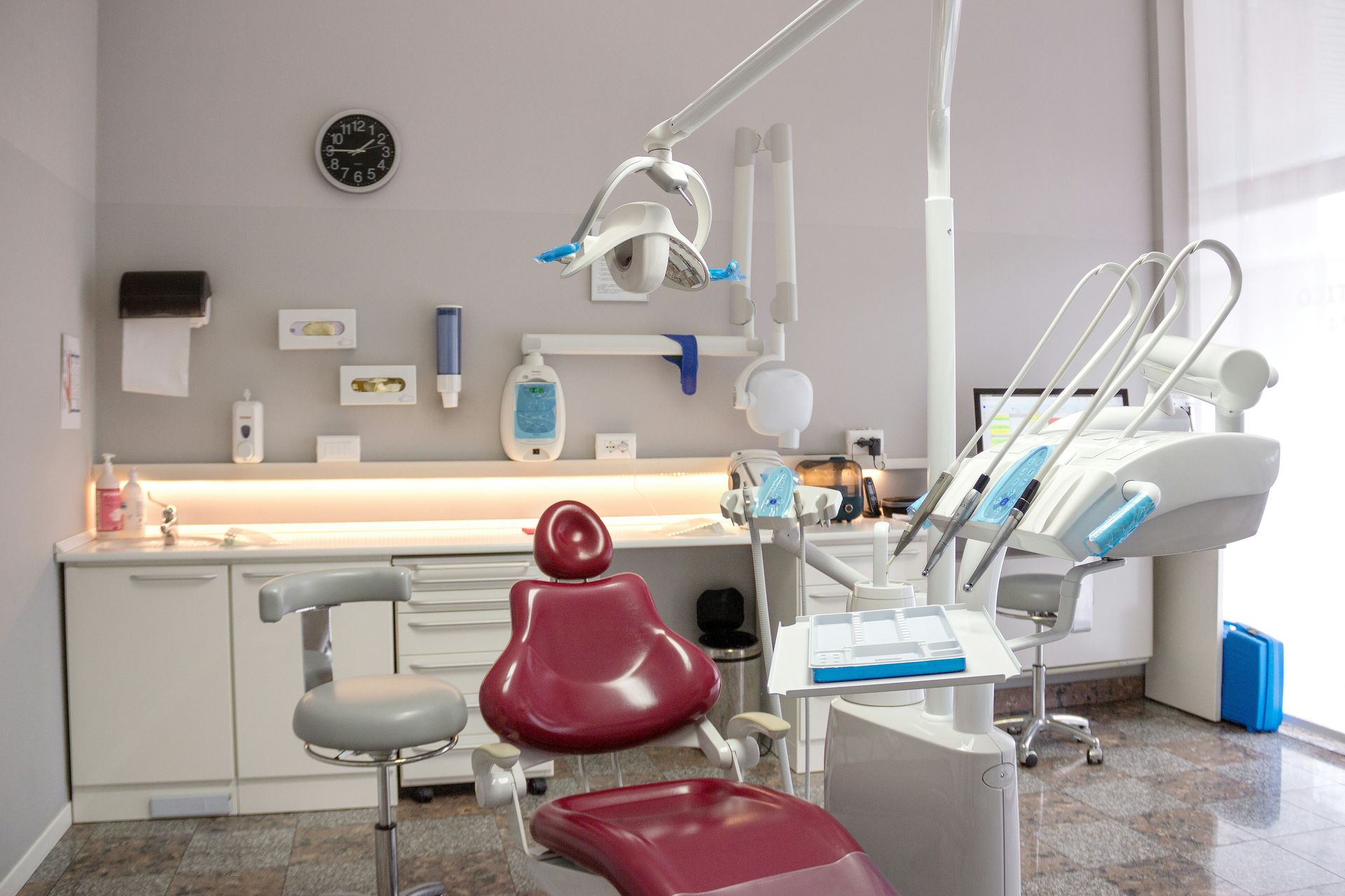 A dental office interior with a patient chair, dental equipment, and cabinets against a light gray wall.