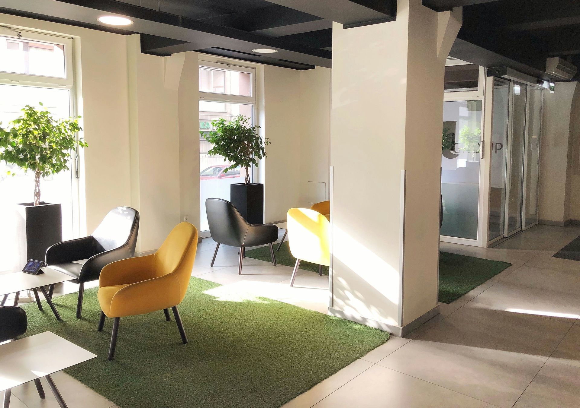 Interior waiting area with yellow and black chairs on green carpet, trees, and glass doors.