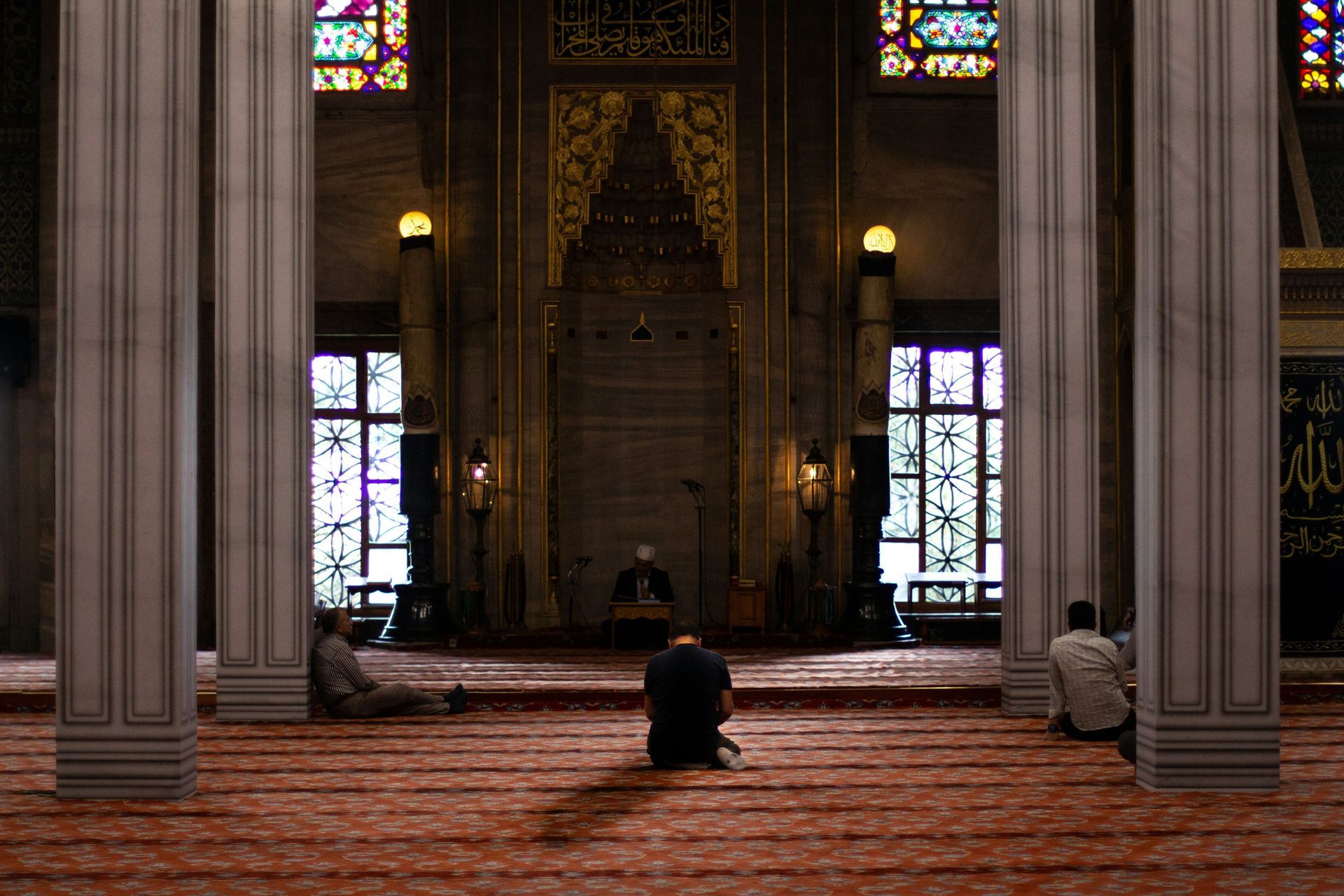 Inside a mosque, individuals kneeling in prayer, facing the ornate mihrab, with large columns and stained glass windows.