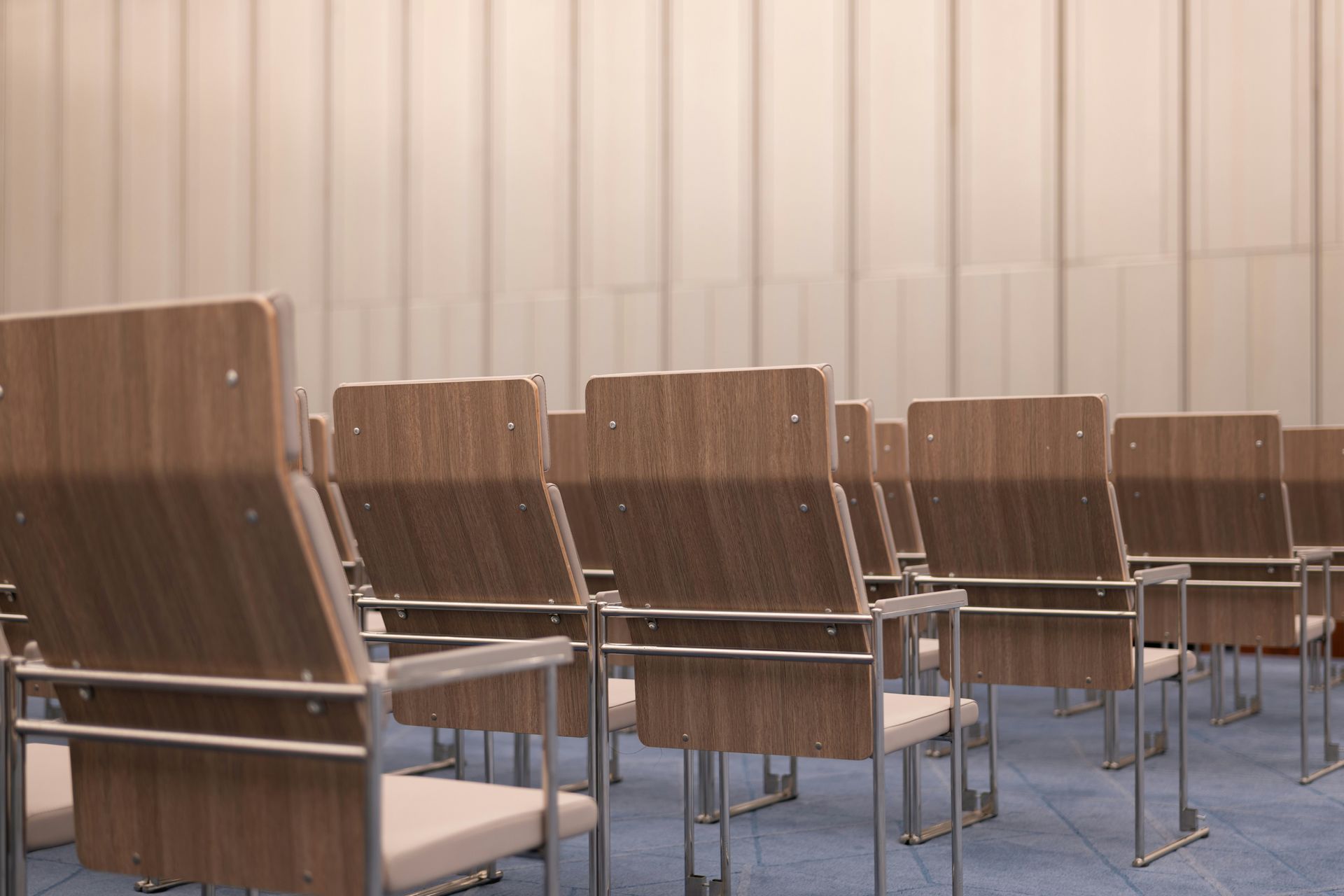 Empty rows of wooden chairs in a conference room, facing away from the viewer; neutral-toned wall in background.