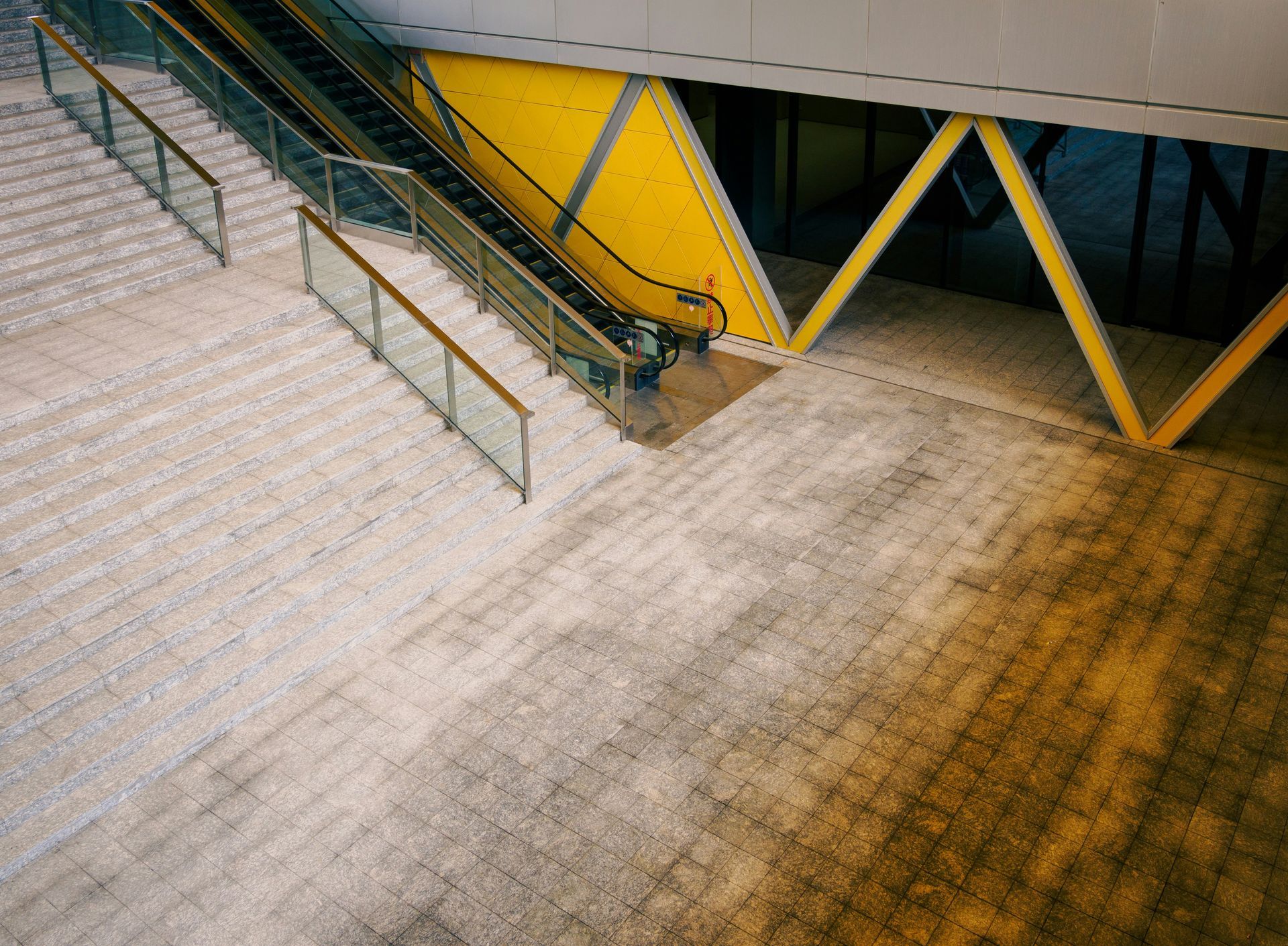 Stairs and an escalator lead up into a modern building with a yellow accent wall and support beams.