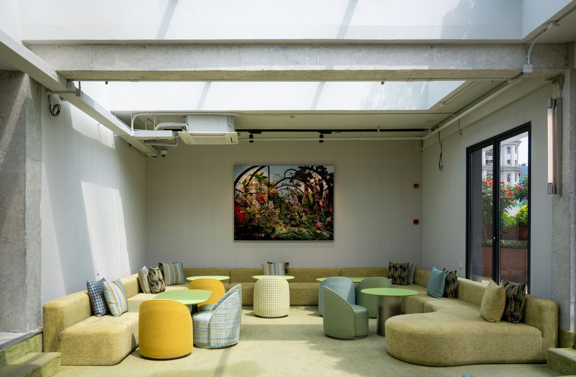 A light-filled lounge with couches, tables, and art. The room has a skylight and a view of outside plants.
