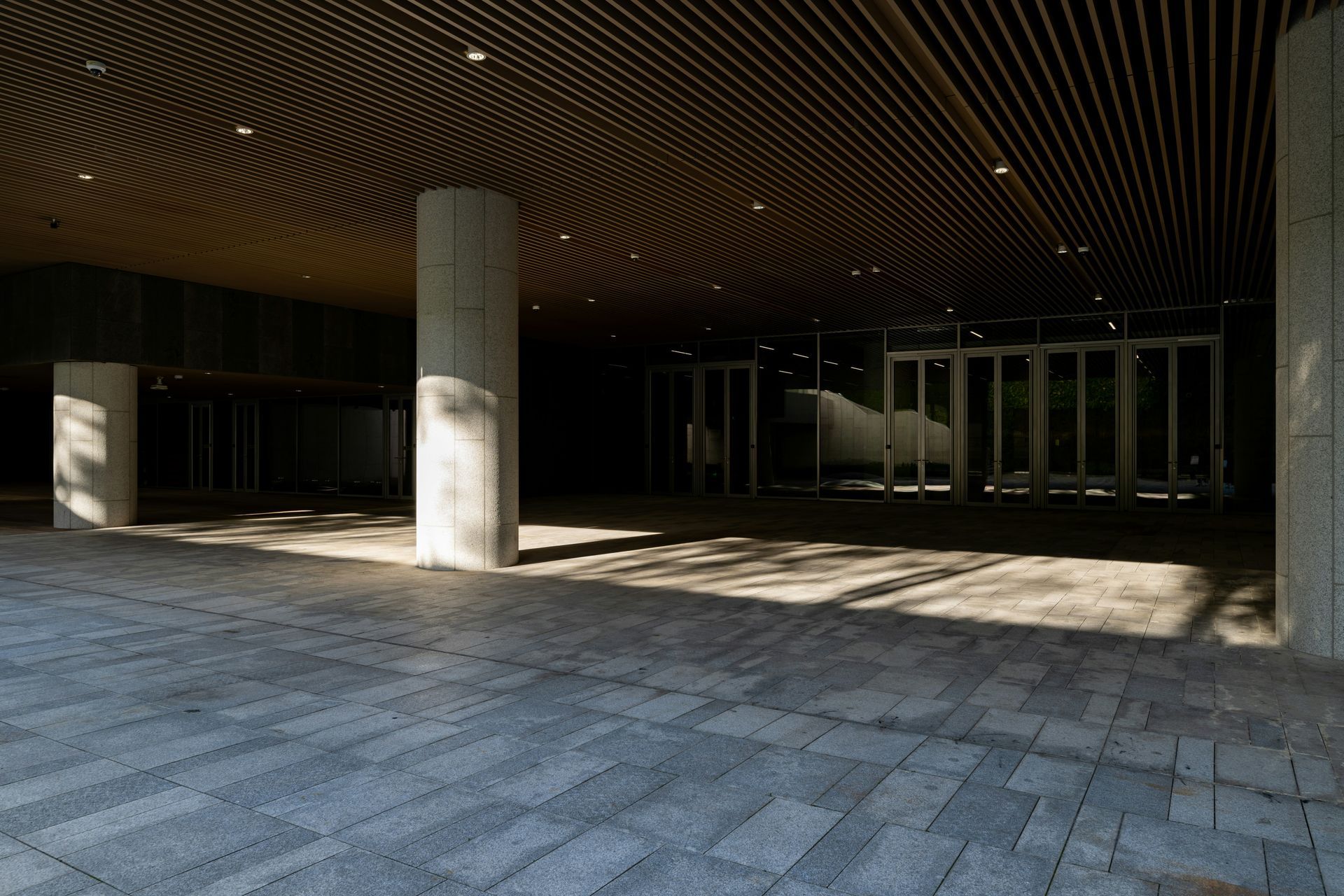 A modern building entrance with stone flooring, concrete columns, and a wooden slatted ceiling; sunlight streams in.