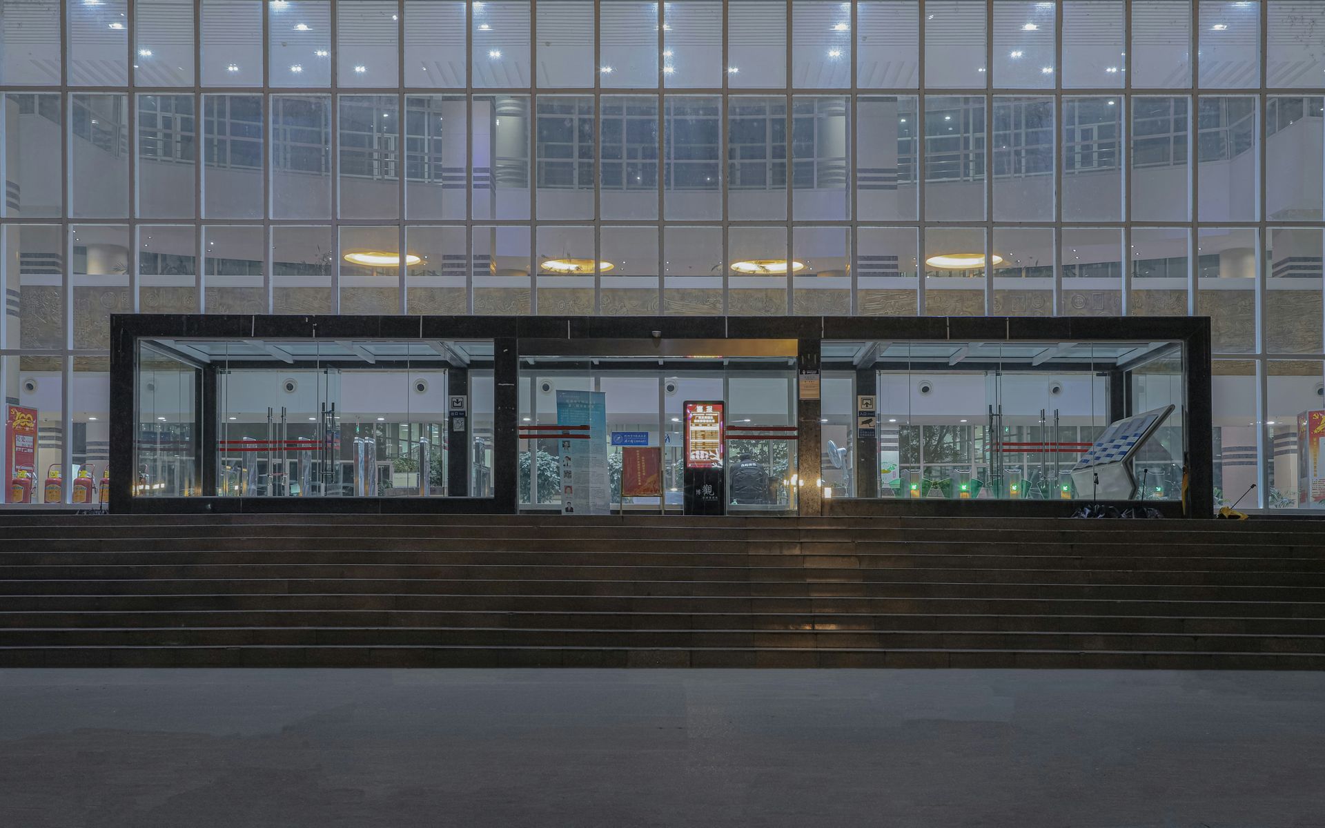 Large glass building entrance with steps leading to it. Lit windows reflecting lights.