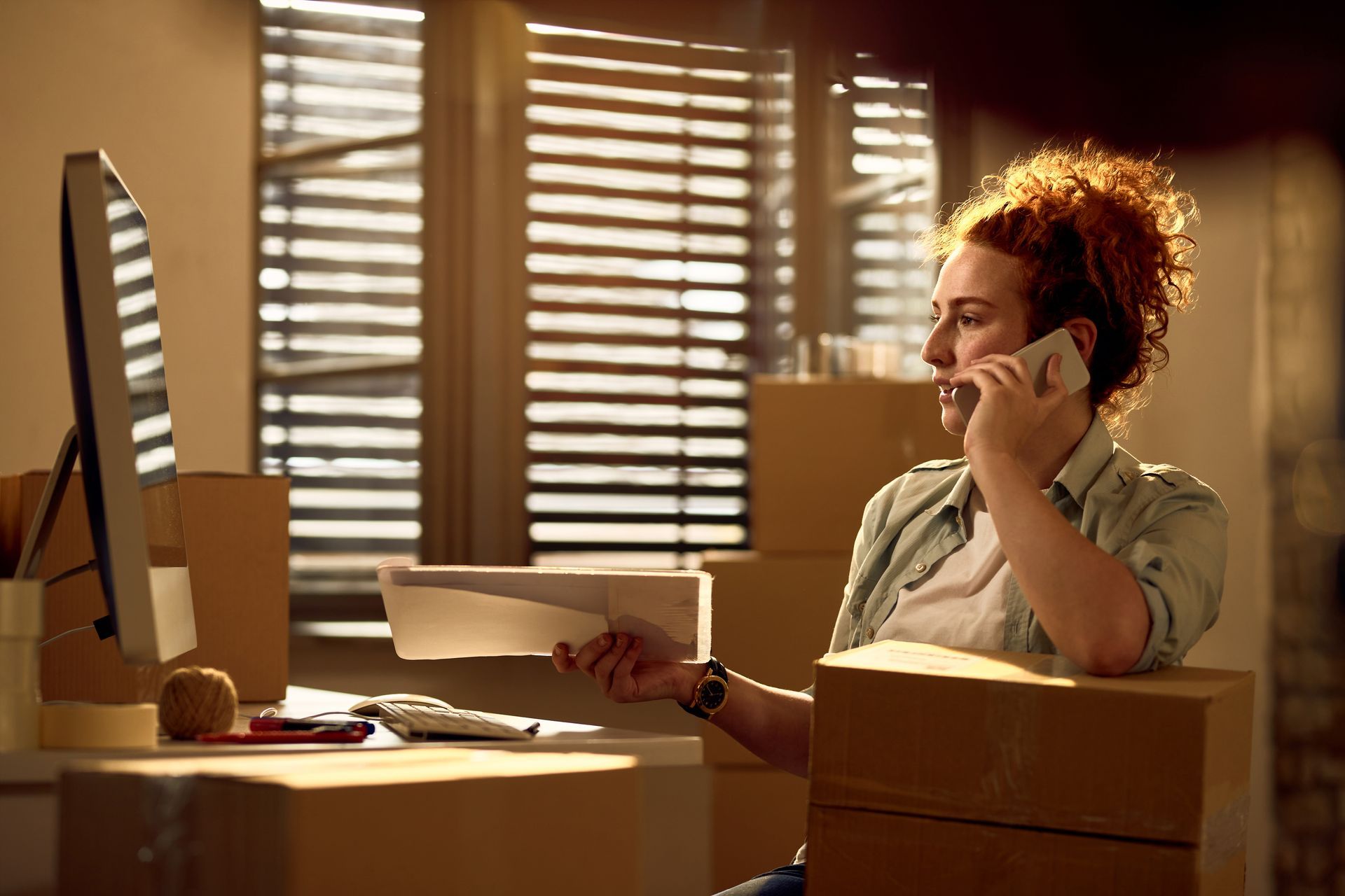 Woman on phone, holding paperwork, surrounded by boxes near computer and window with sunlight.