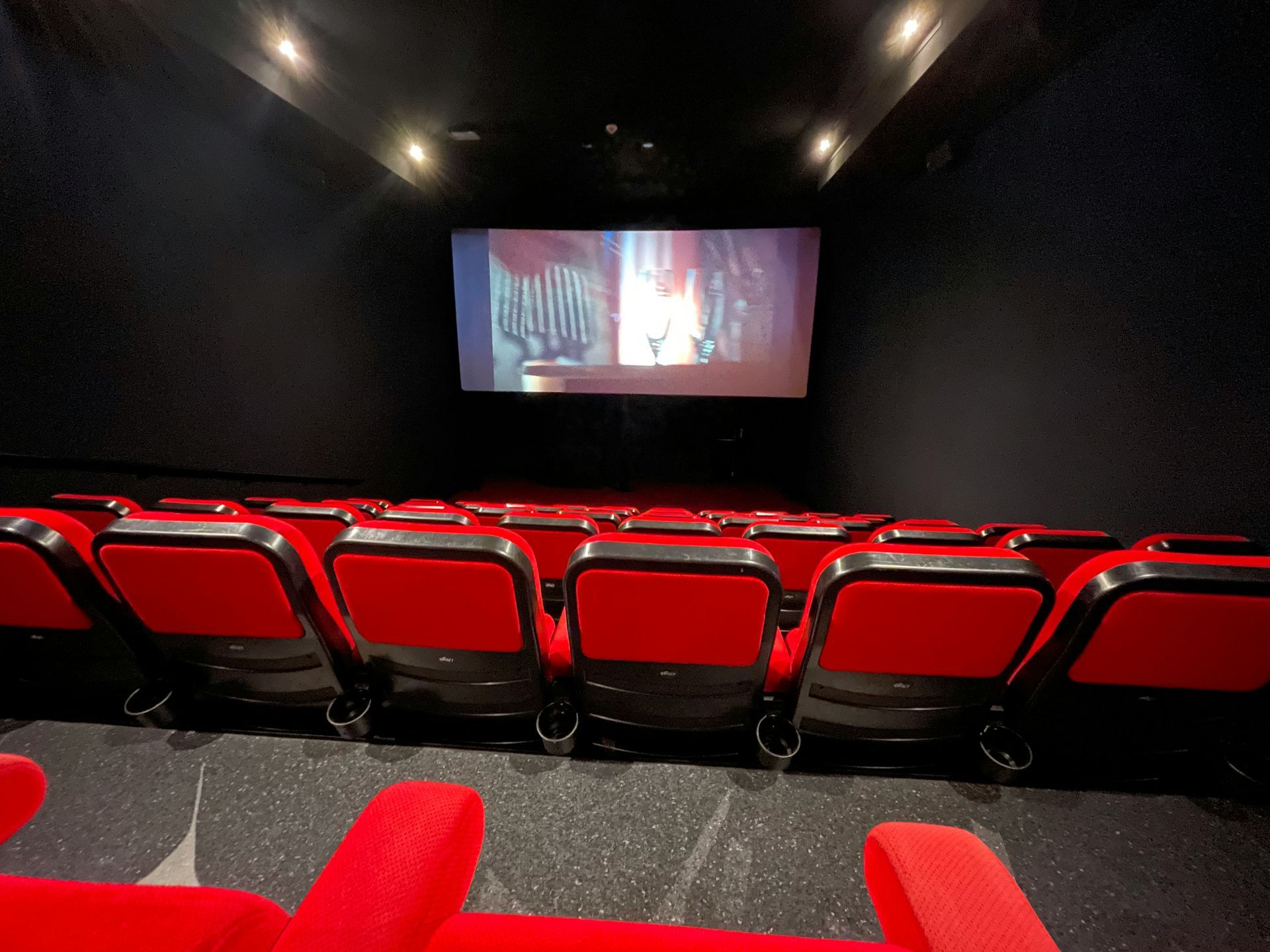 Empty red theater seats face a movie screen. Dark walls, dim lights.