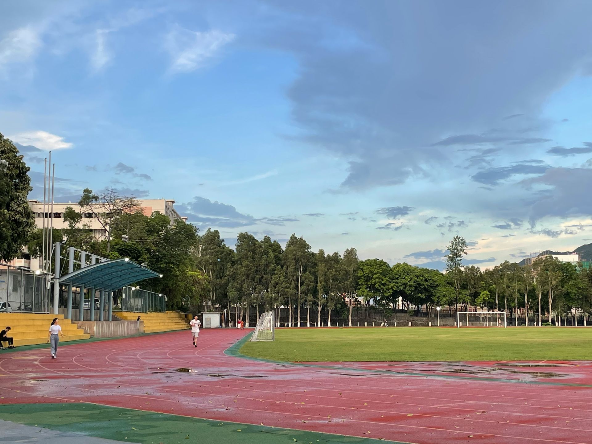 Red track, green field, and trees under a cloudy sky. Two people are jogging.