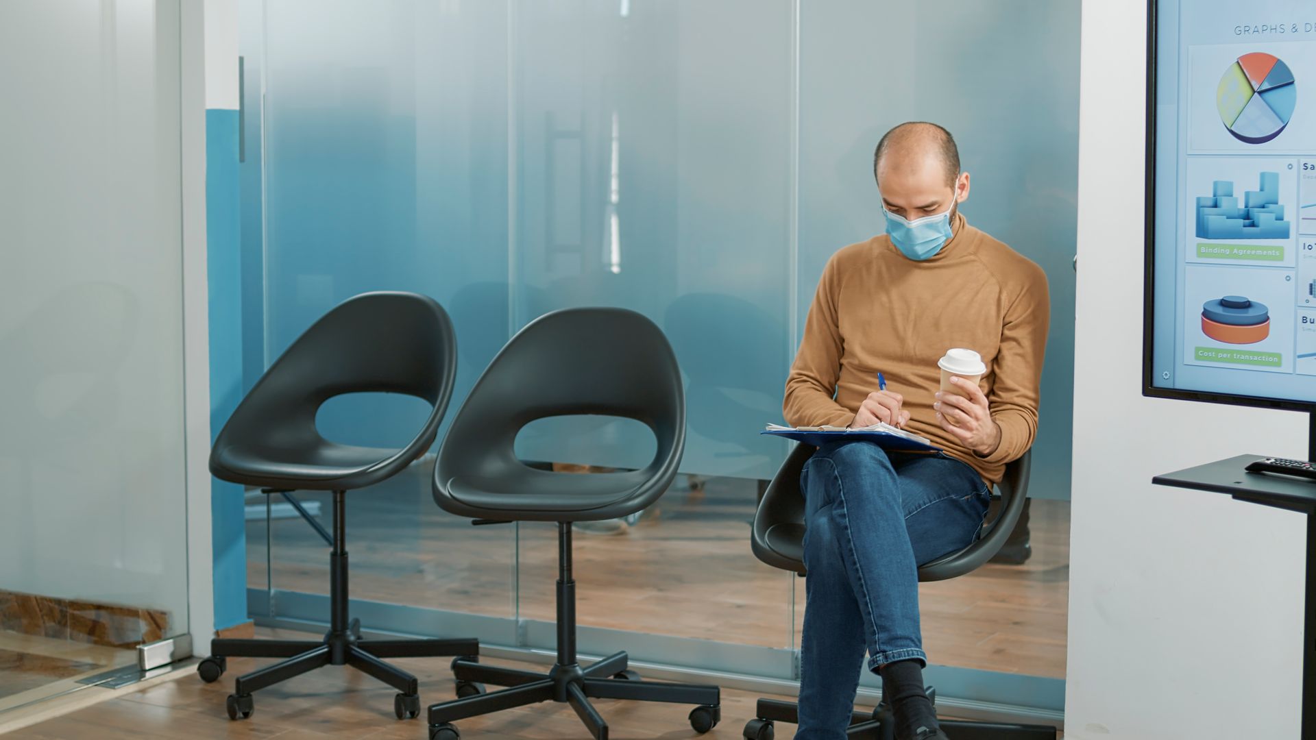 Man in mask, sitting in waiting area, writing on pad, holding coffee.