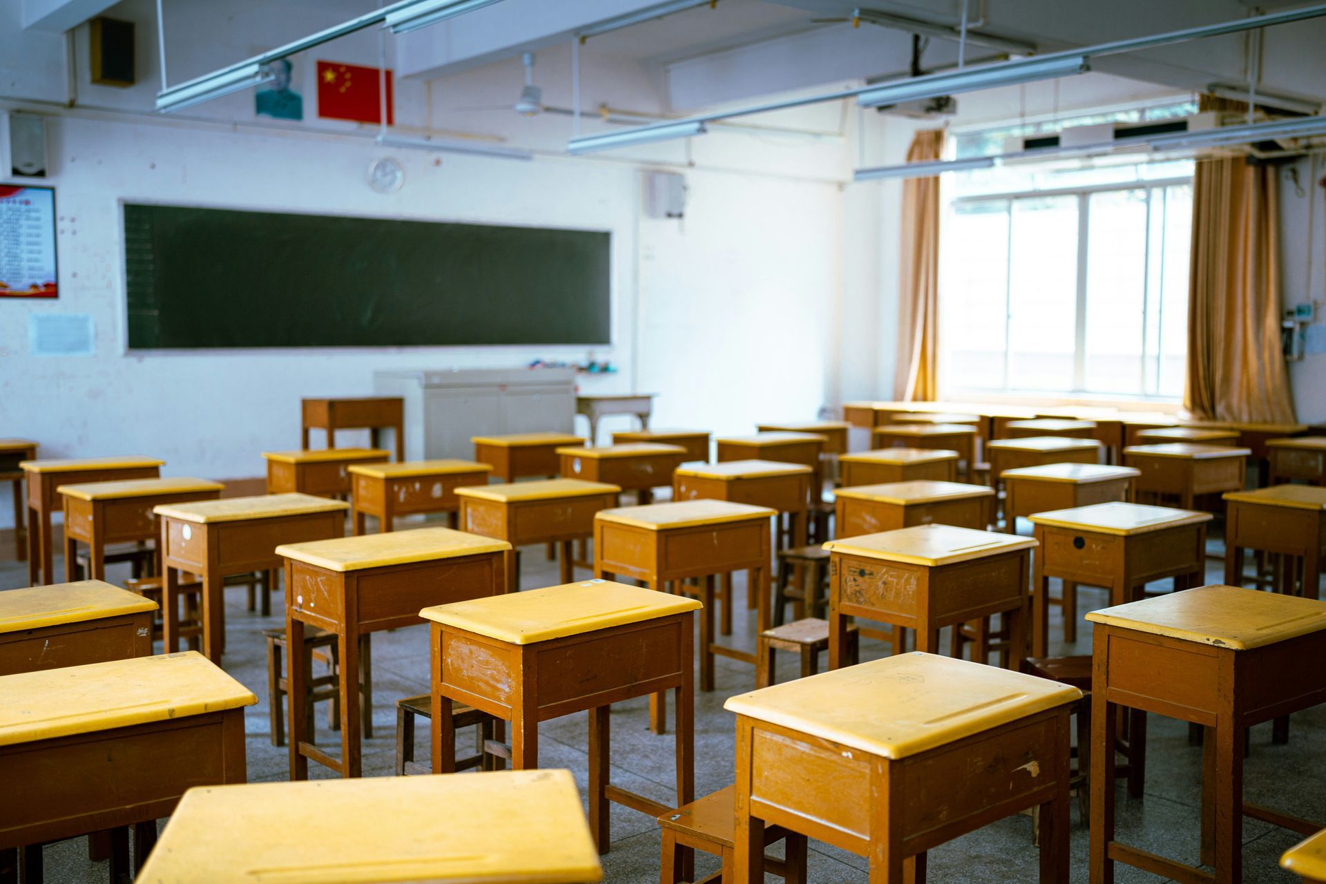 Empty classroom with rows of wooden desks facing a chalkboard, a window with curtains, and a Chinese flag.