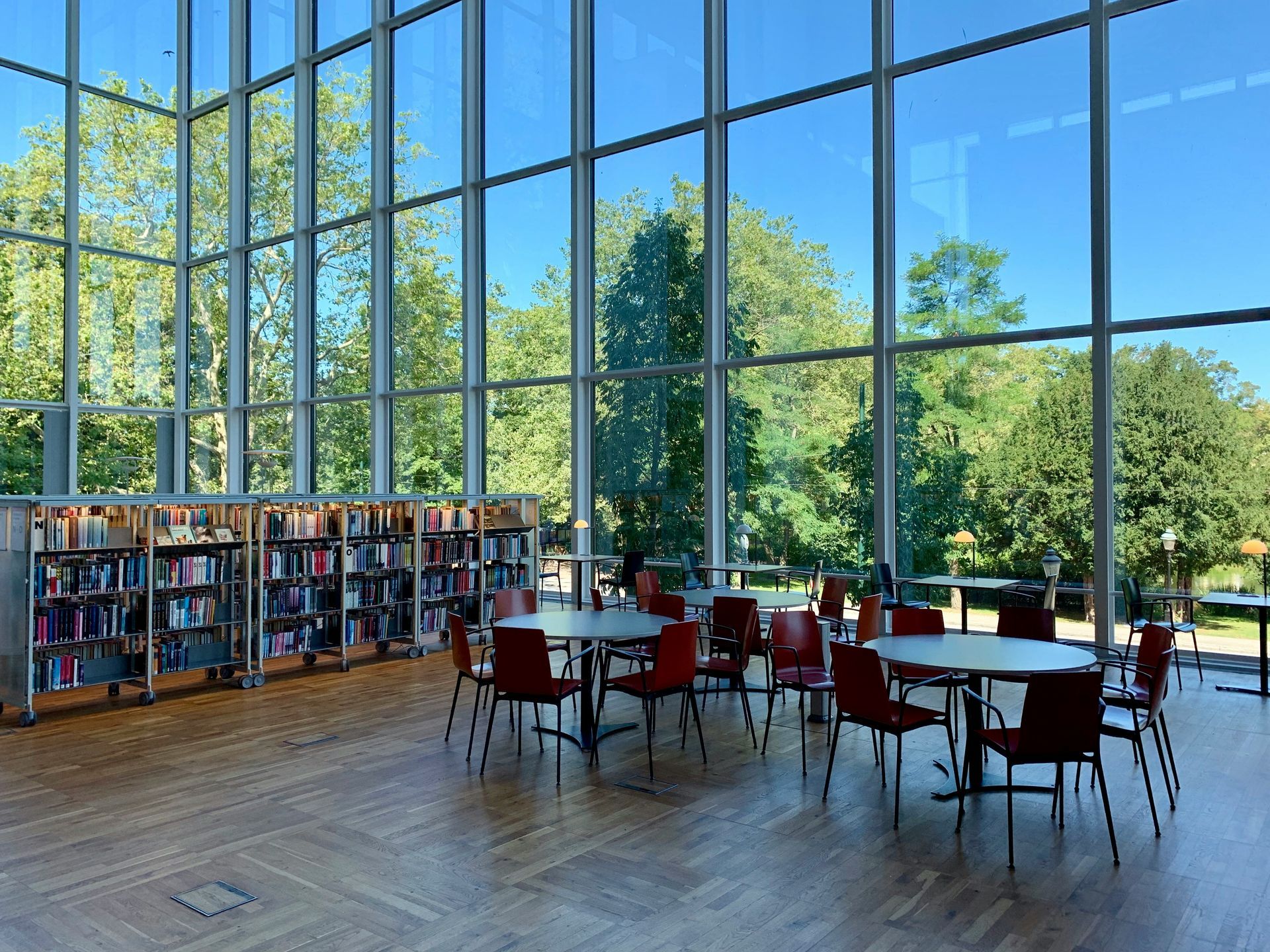 Library interior with floor-to-ceiling windows, bookshelves, round tables, and red chairs overlooking a tree-filled park.