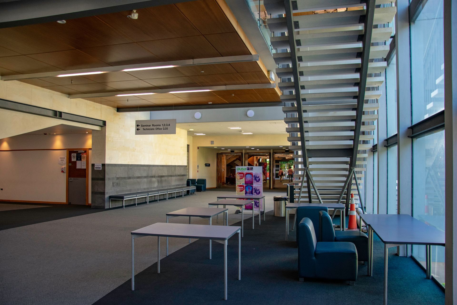 Interior hallway with stairs, tables, and seating by large windows.