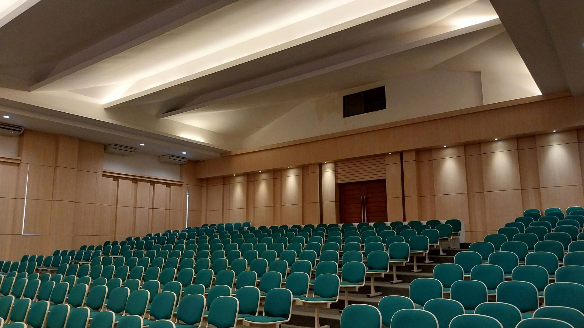 An auditorium with tiered seating filled with teal chairs, beige walls, and a stage.