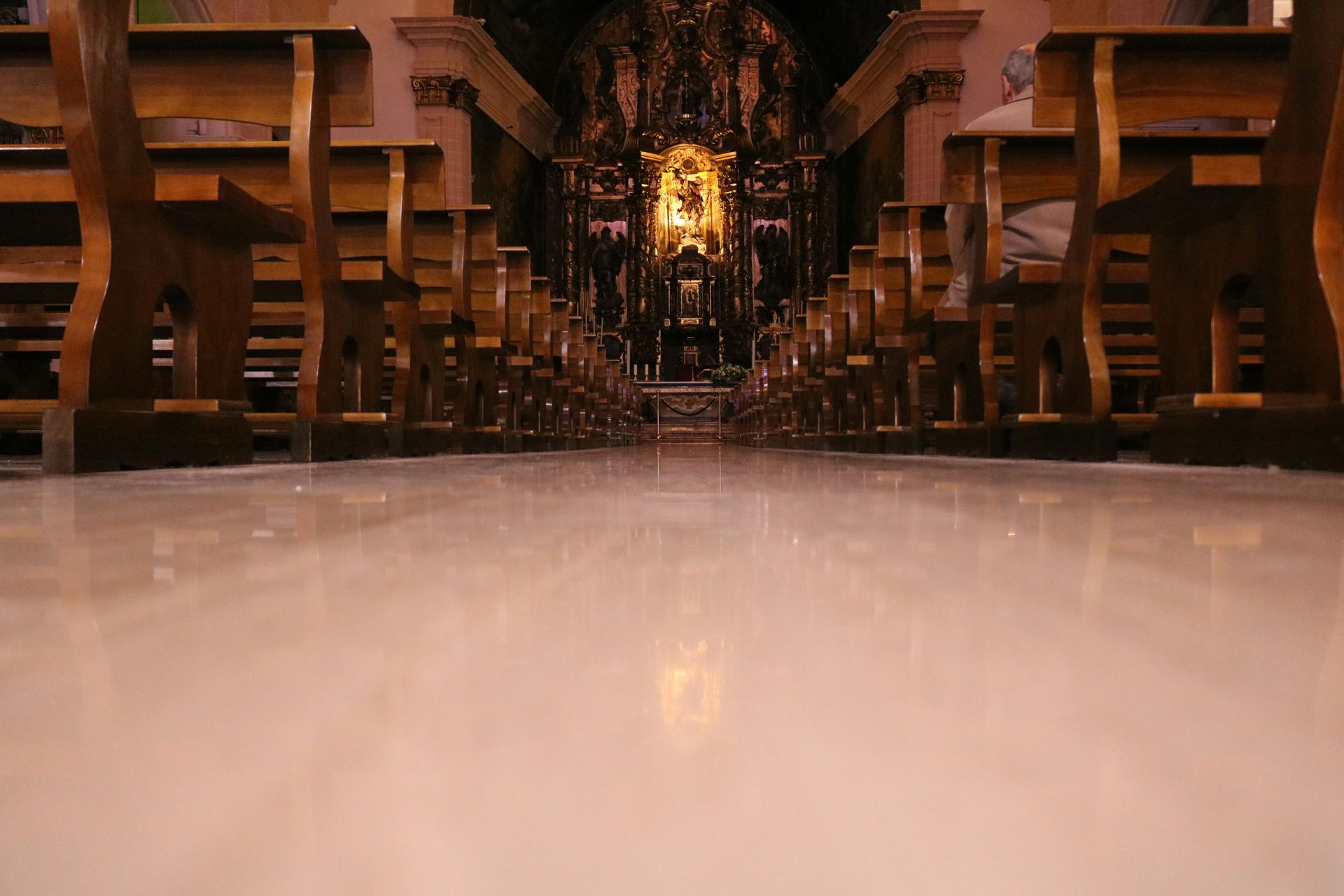 Interior of a church with rows of wooden pews, leading to a golden altar. Polished floor reflects the space.
