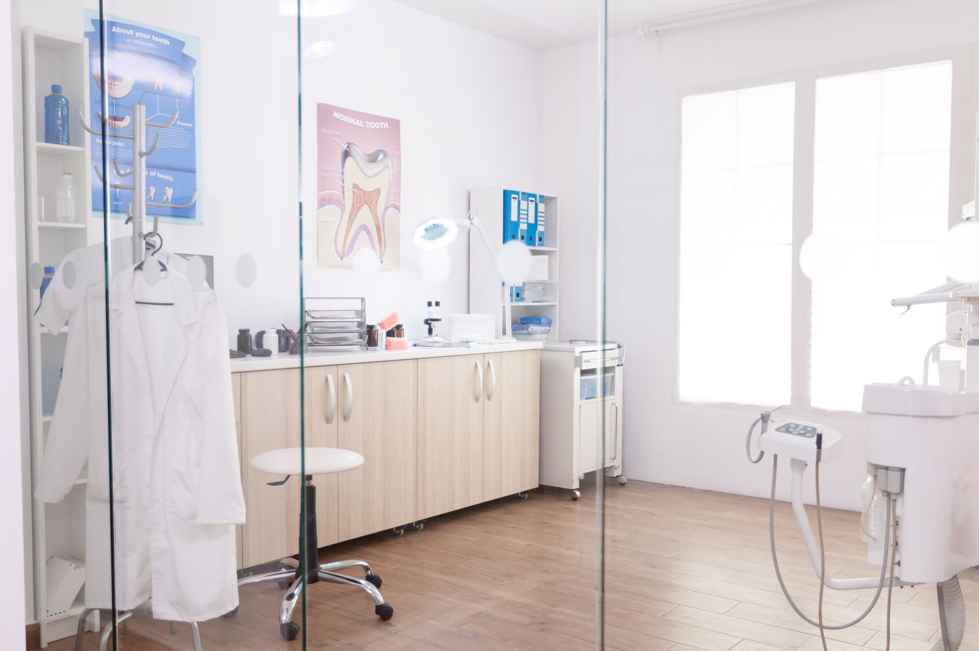 Dental office with exam room visible through glass wall. Equipment, cabinets, posters, and medical coat are present.