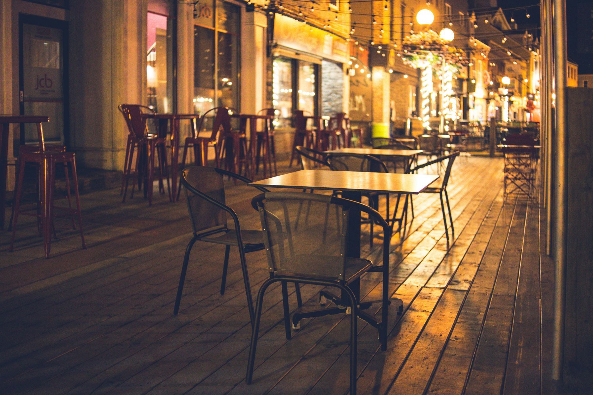 Outdoor restaurant seating, tables and chairs on a wooden deck at night, lit by overhead lights.