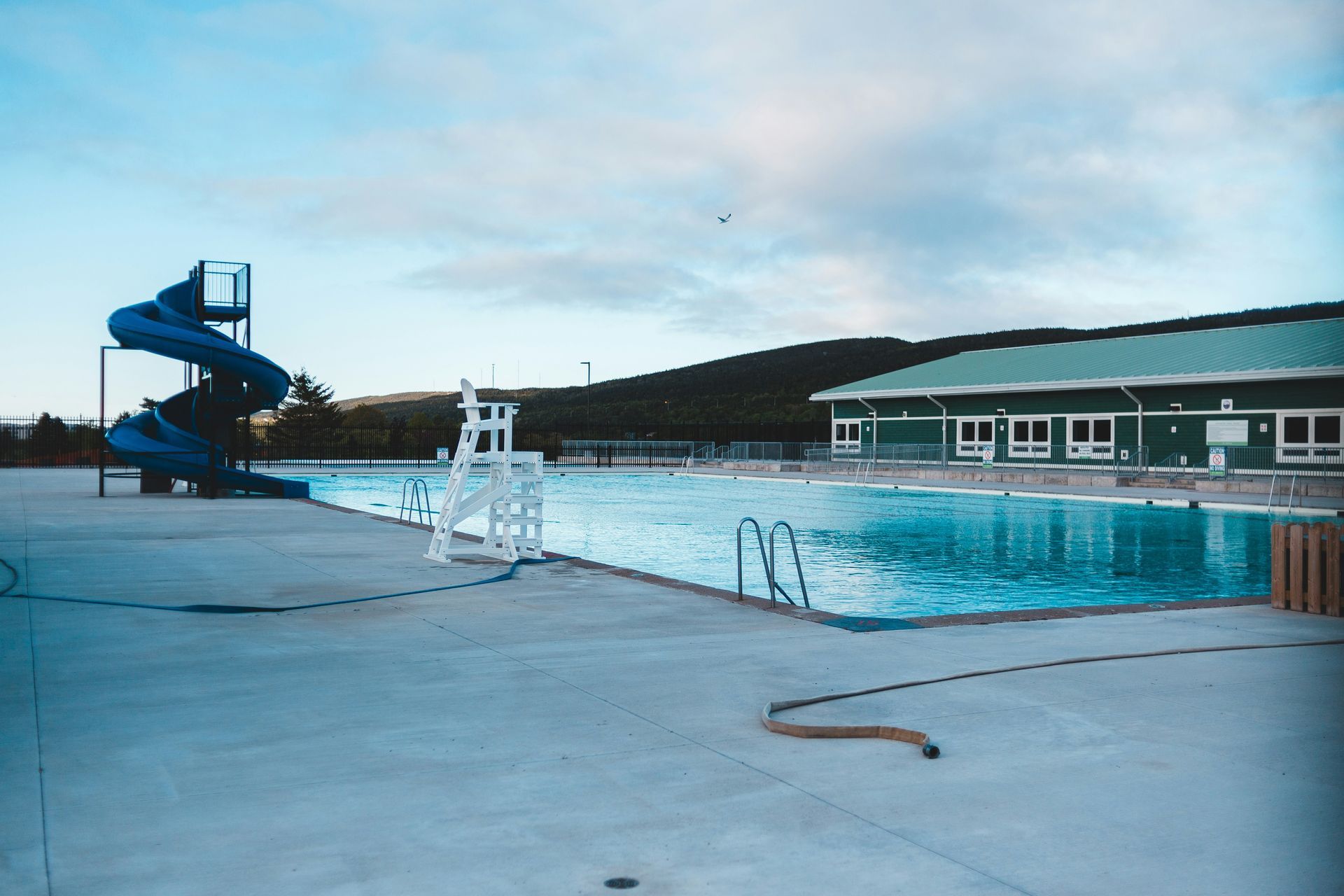 Blue swimming pool with a slide, lifeguard stand, and building under a cloudy sky.