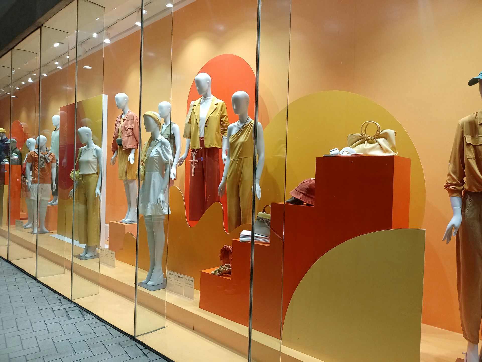 Storefront display with mannequins in orange and yellow clothing; geometric backdrop.