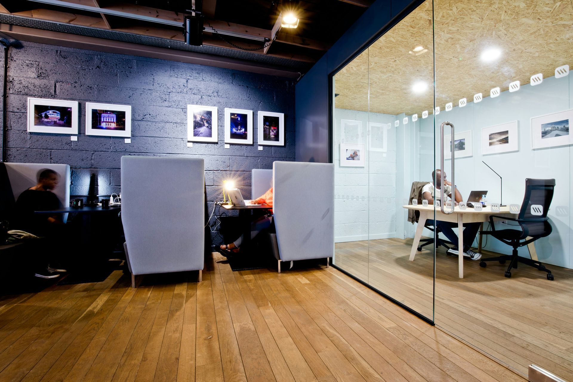 Interior view of an office space with wooden floors, workstations, and a glass-walled office. People work at desks.