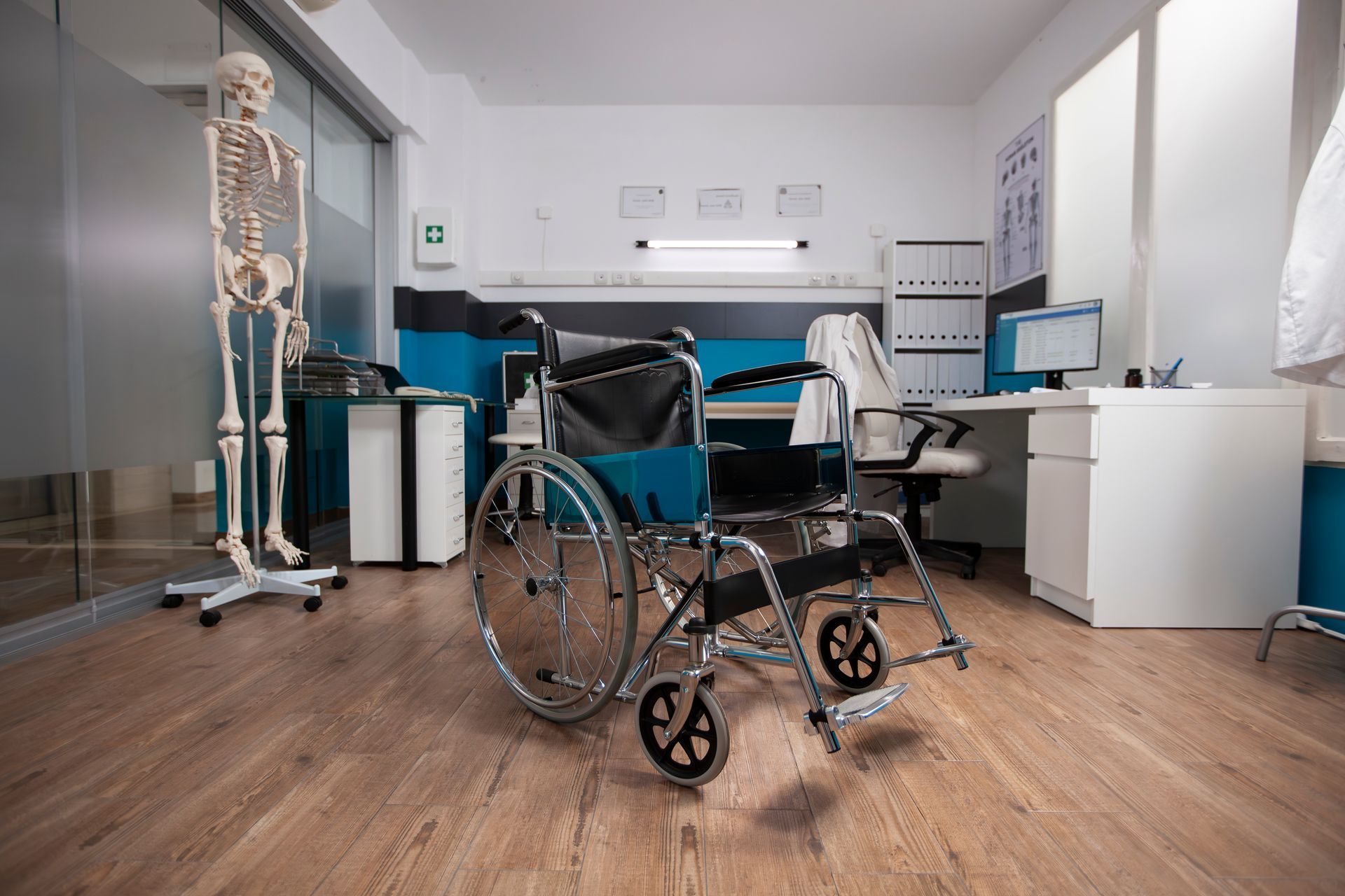 Wheelchair in a doctor's office with a skeleton, desk with computer, and medical chart.