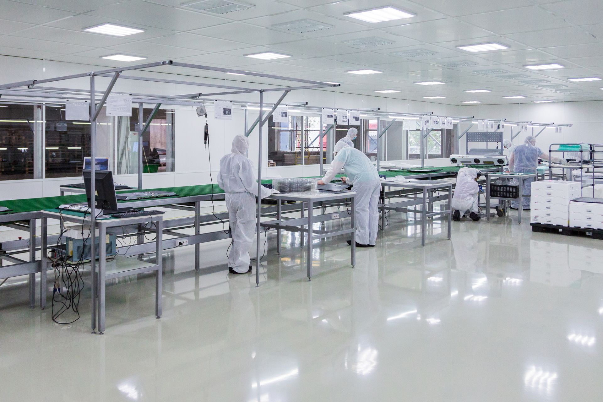 Workers in white protective suits assemble products in a cleanroom. Shiny surfaces and overhead conveyors.