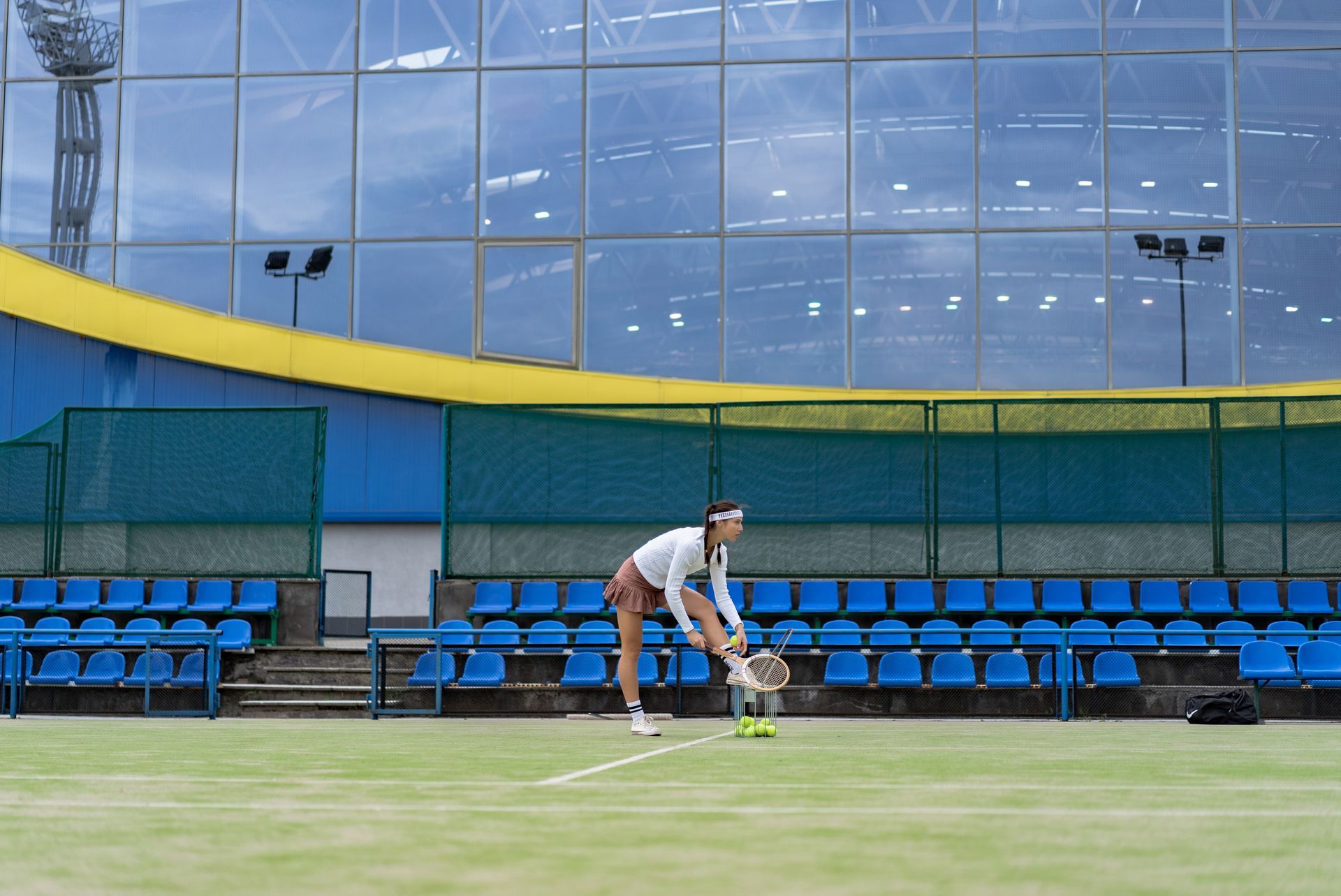 Woman stretching to pick up tennis balls on a green court in front of blue stadium seats.