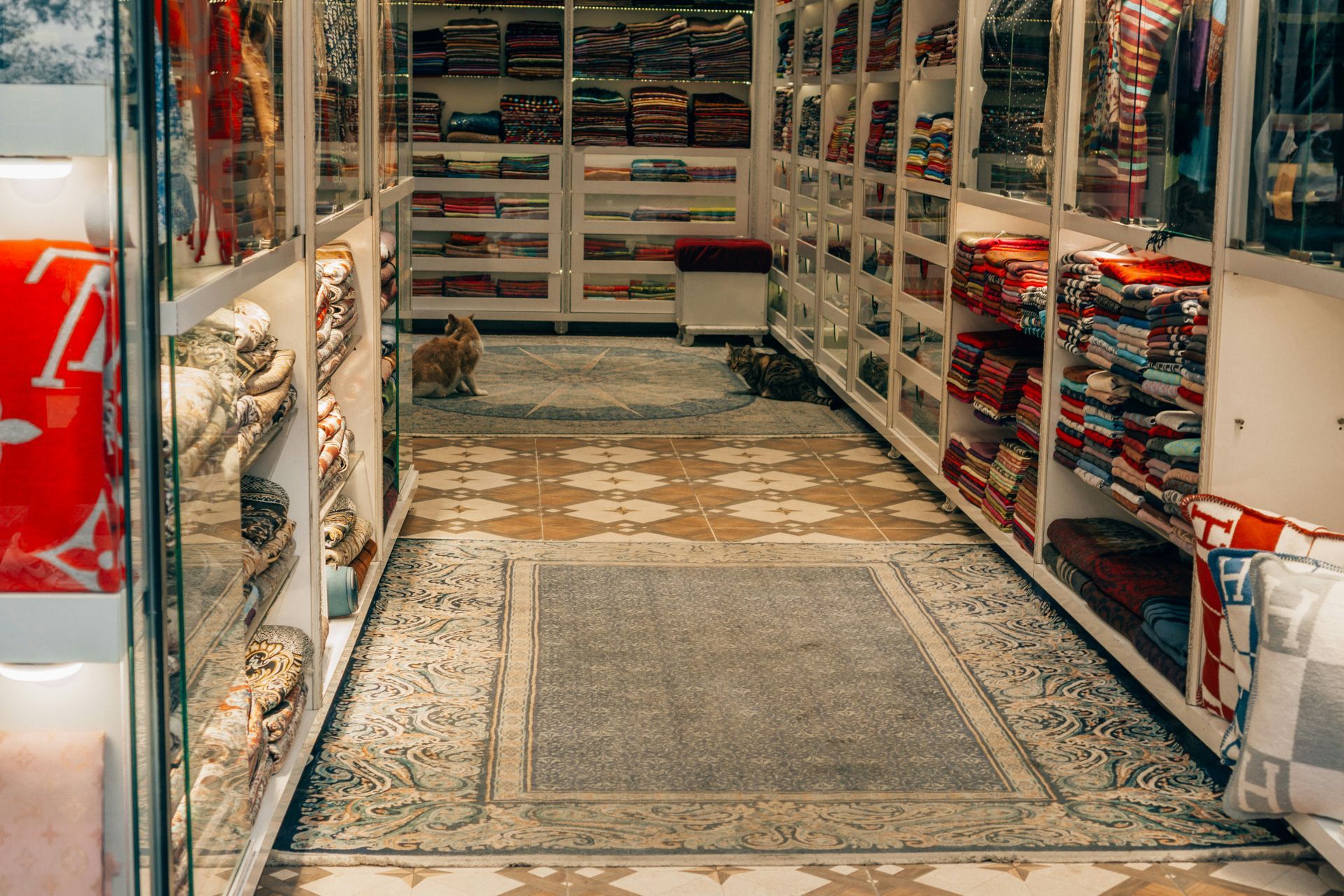 Narrow store aisle with shelves of colorful textiles; two cats rest on patterned rugs.
