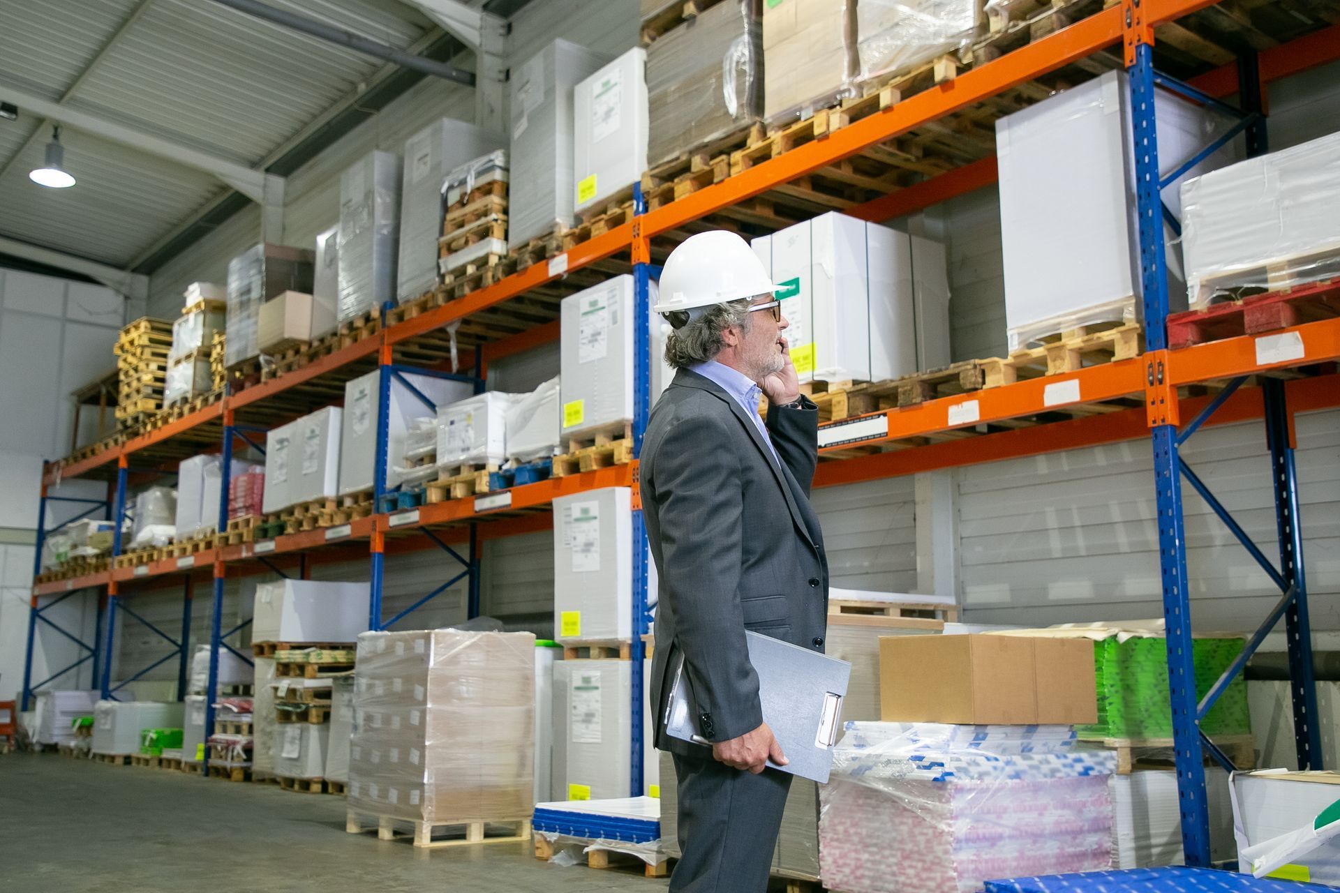 Man in suit and hard hat on phone in a warehouse with shelving packed with boxes and materials.