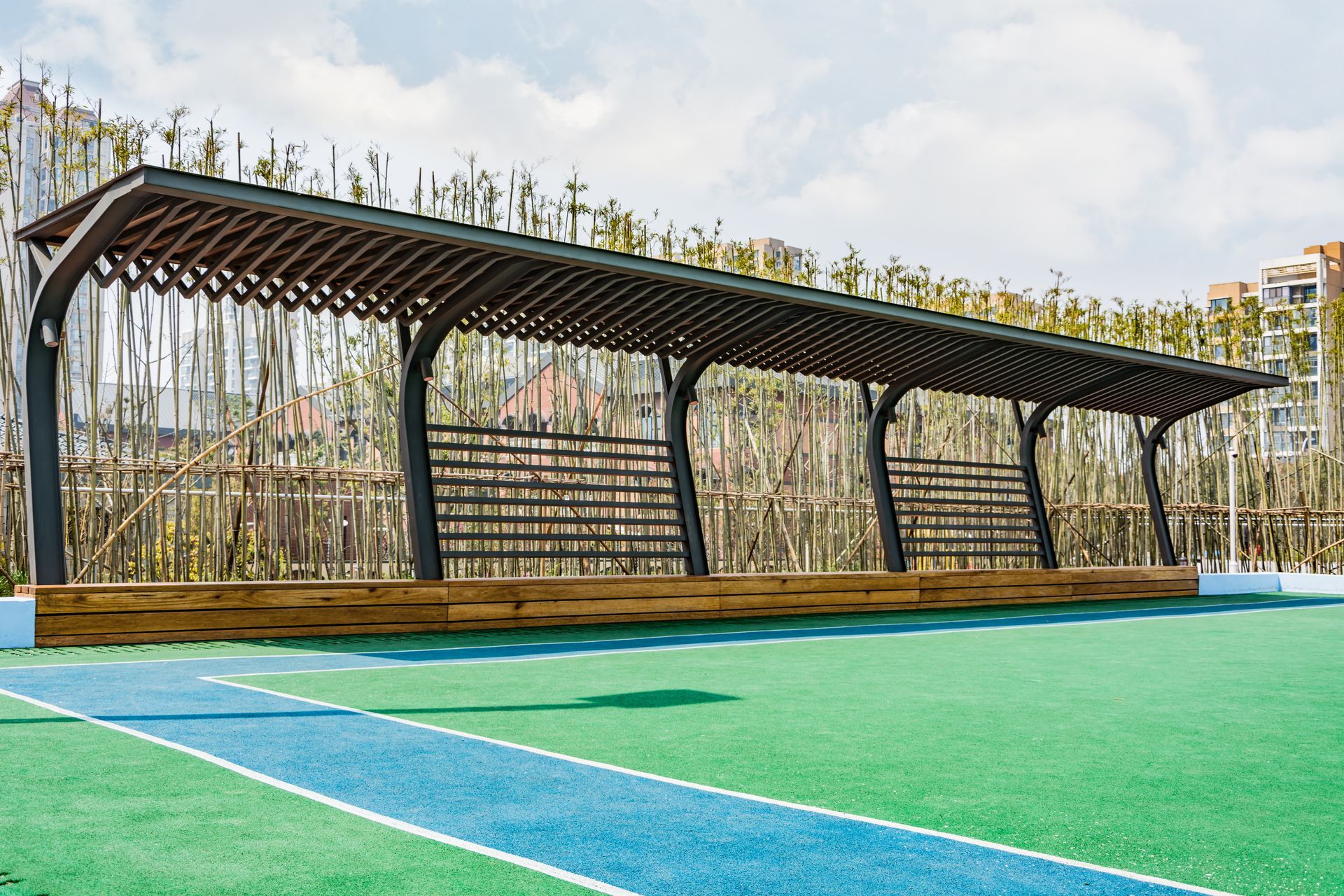 A covered seating area with a wooden bench, on a green surface with a blue pathway, against a fence and some foliage.