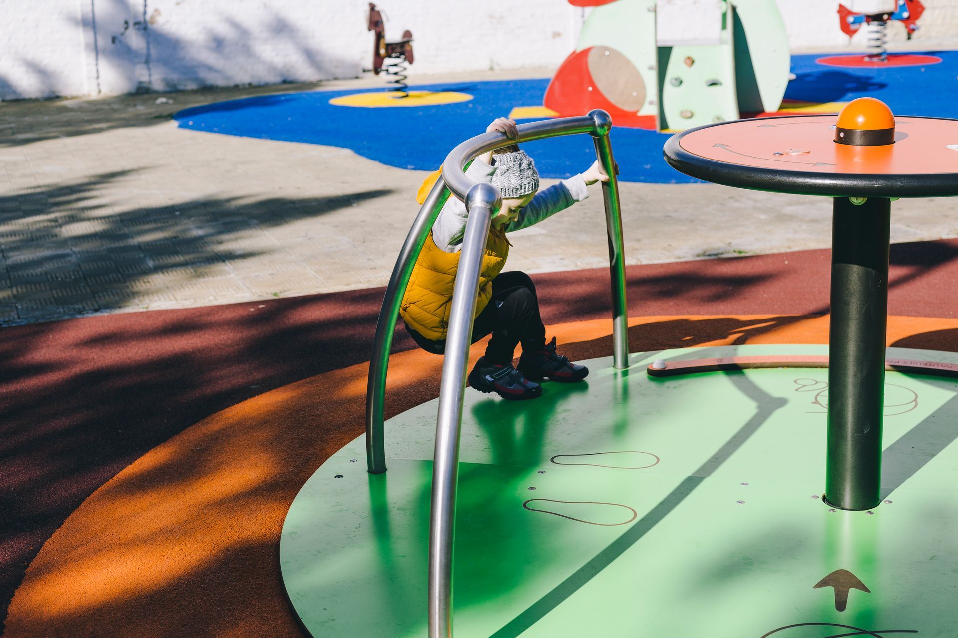 Child playing on a playground merry-go-round, holding onto the bars. Yellow vest.