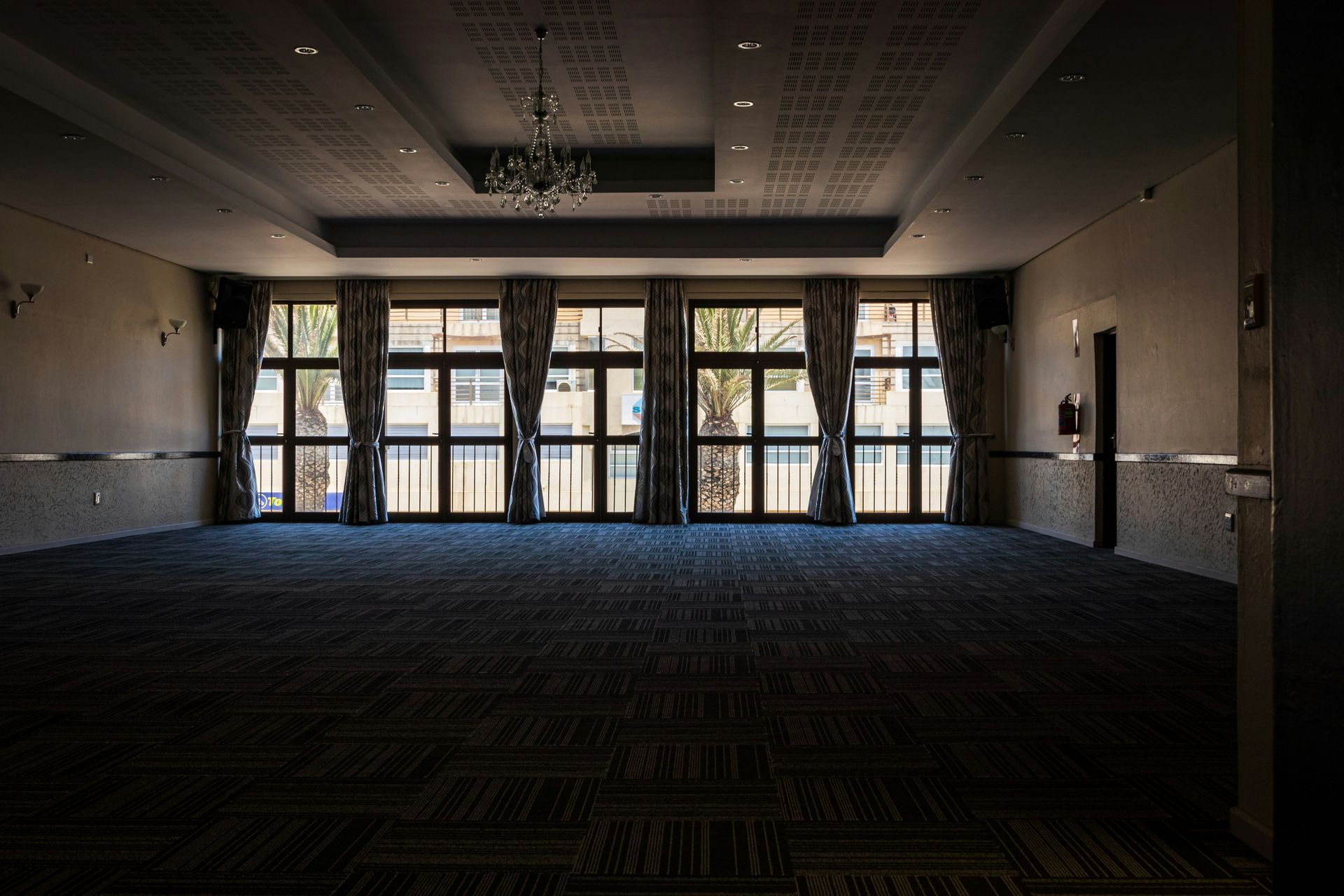 Empty, spacious room with large windows, dark carpet, and a chandelier.