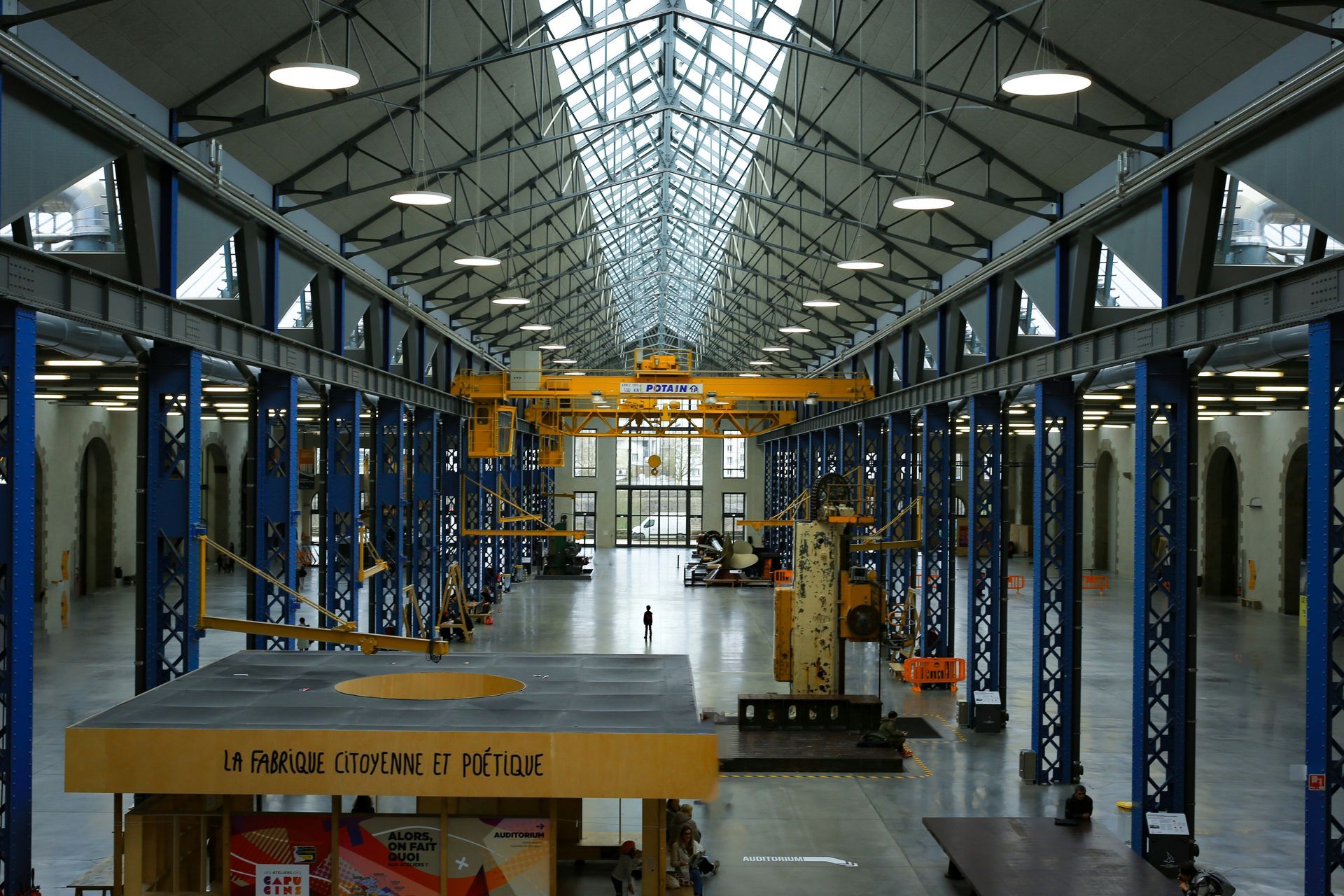 Large industrial building interior with metal beams, overhead cranes, and natural light from a glass roof.