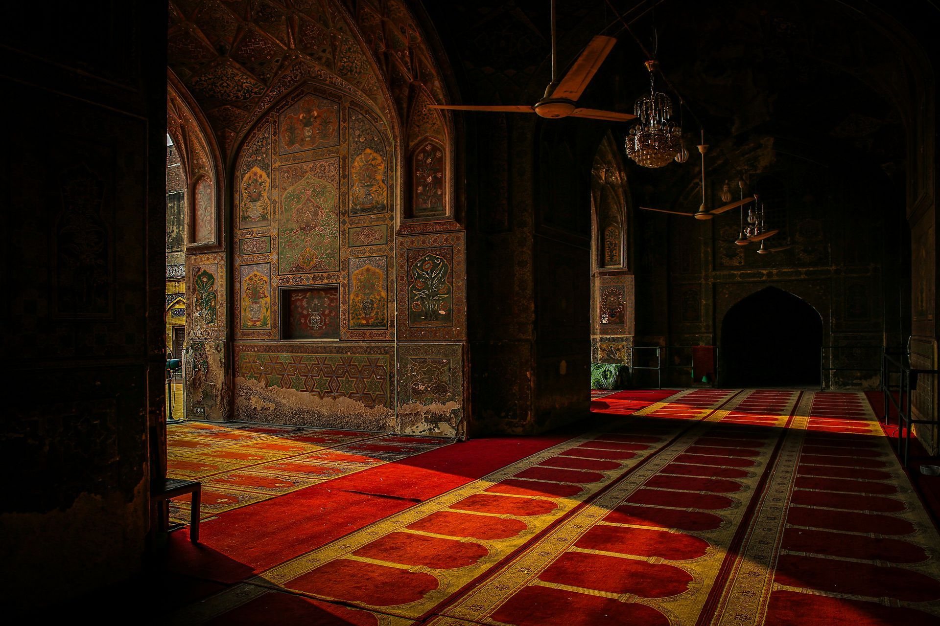 Interior of a mosque with red carpets, arched doorways, ornate walls, and sunlight streaming in.