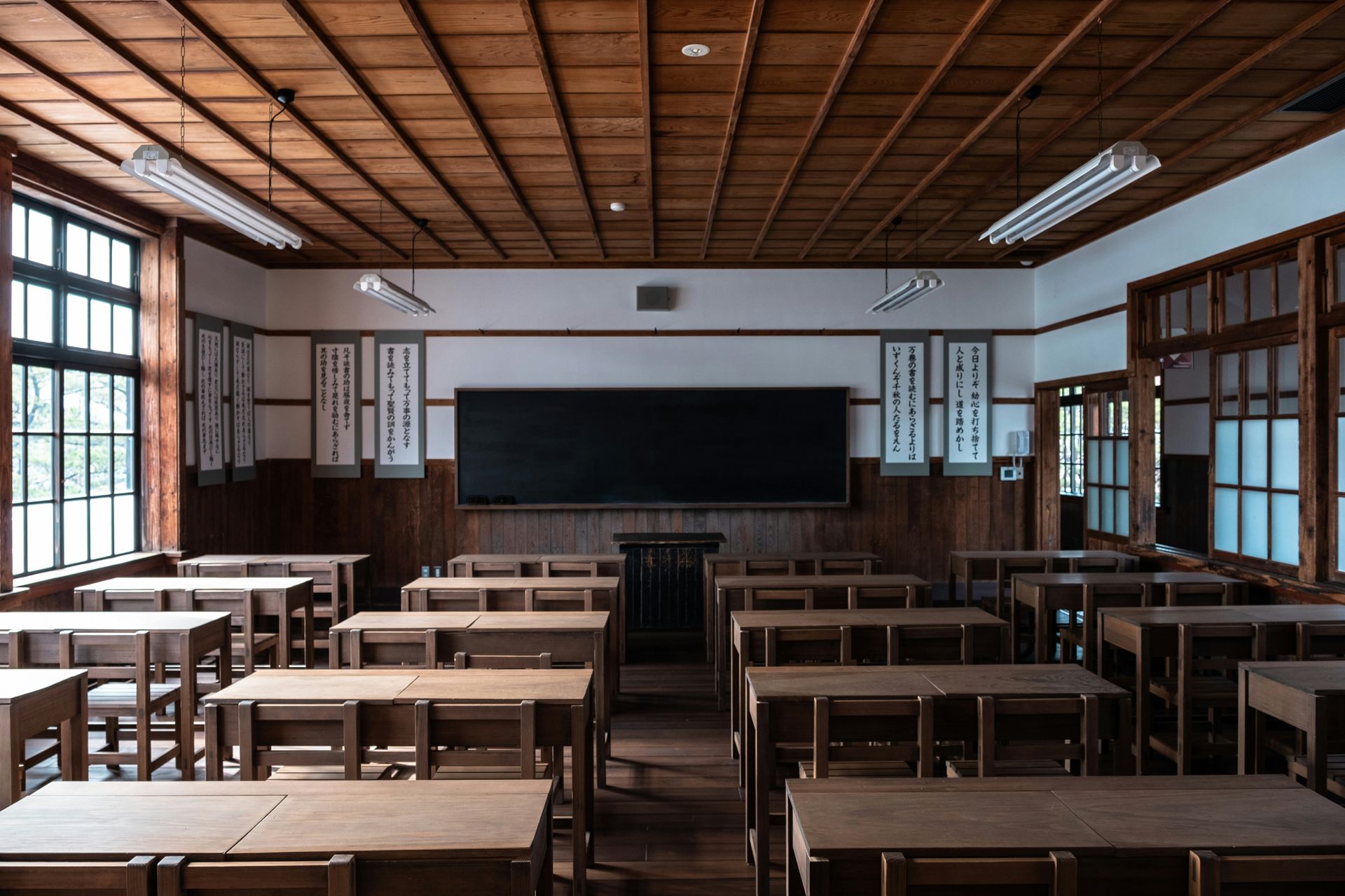 Empty, wood-paneled classroom with rows of desks, a large chalkboard, and large windows.