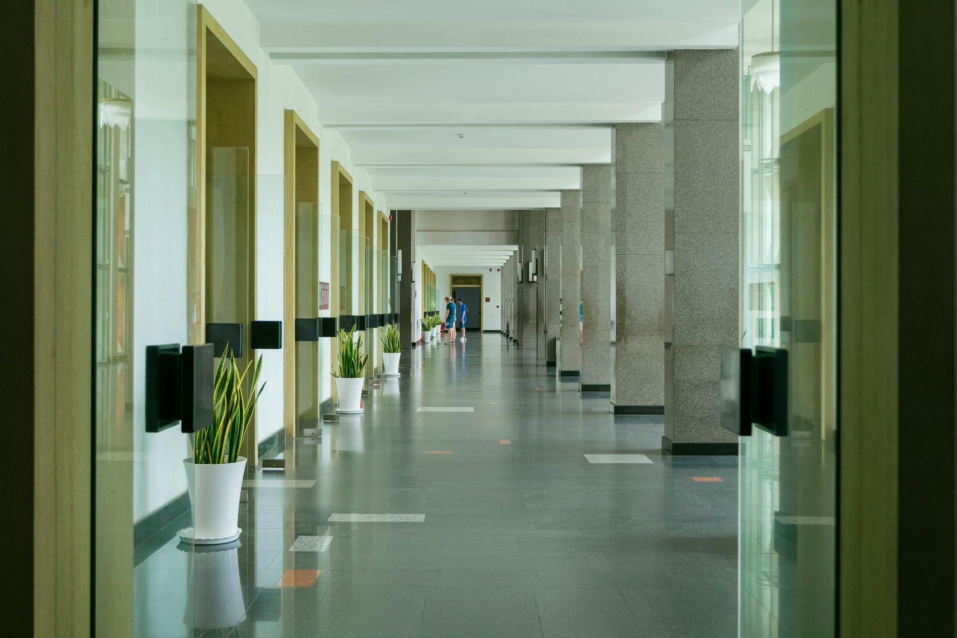 Long hallway with pillars, glass doors, potted plants, and distant figures.