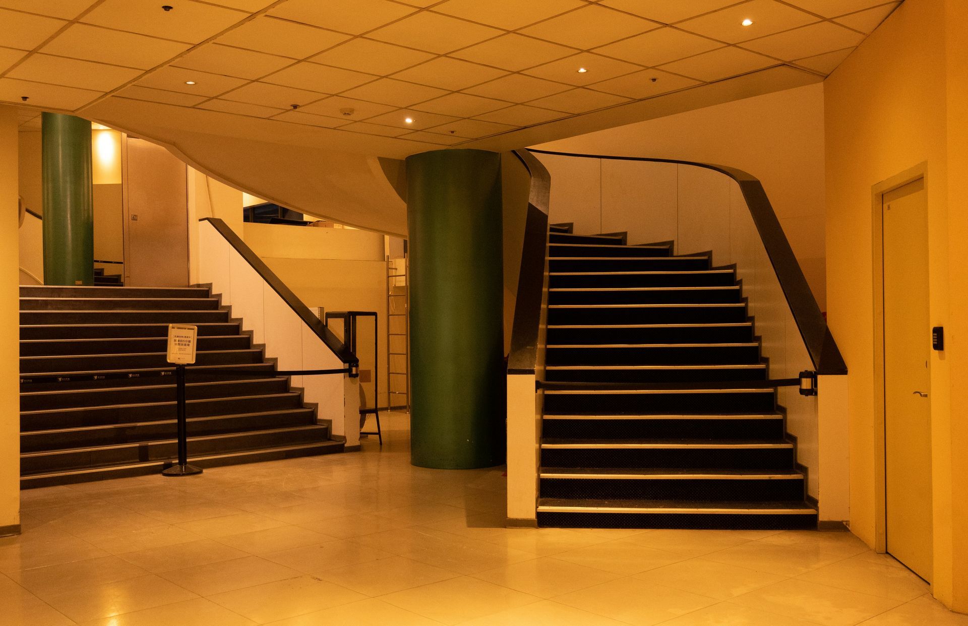Two staircases with dark treads and handrails in a yellow-lit interior. A green pillar is between them.