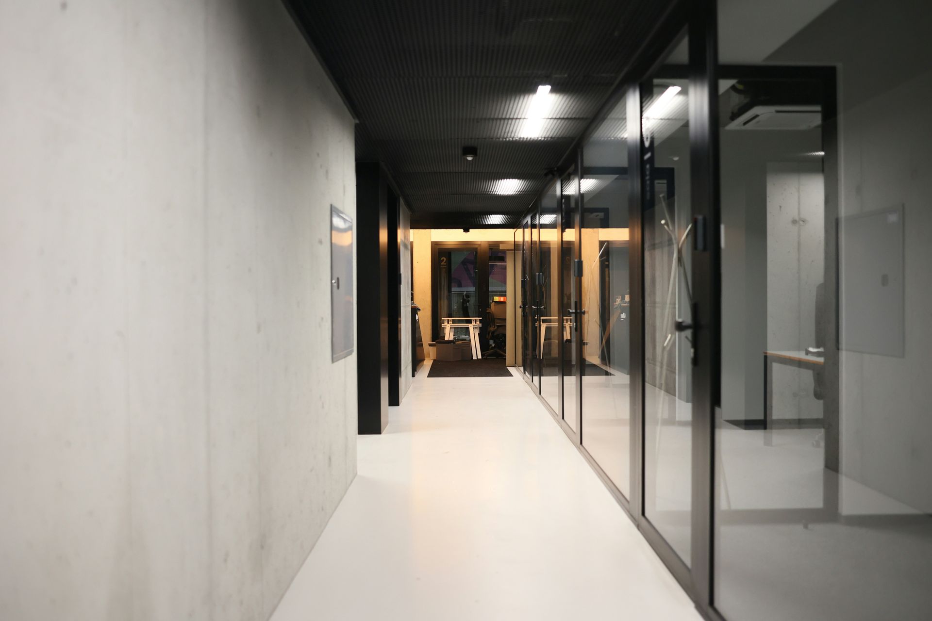 Long hallway with offices on the right side. White floor and walls, black ceiling. Glass doors.