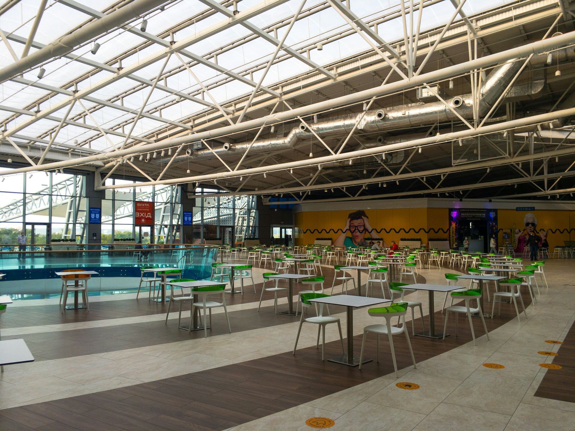 Indoor food court with tables and chairs, adjacent to a swimming pool under a glass roof.