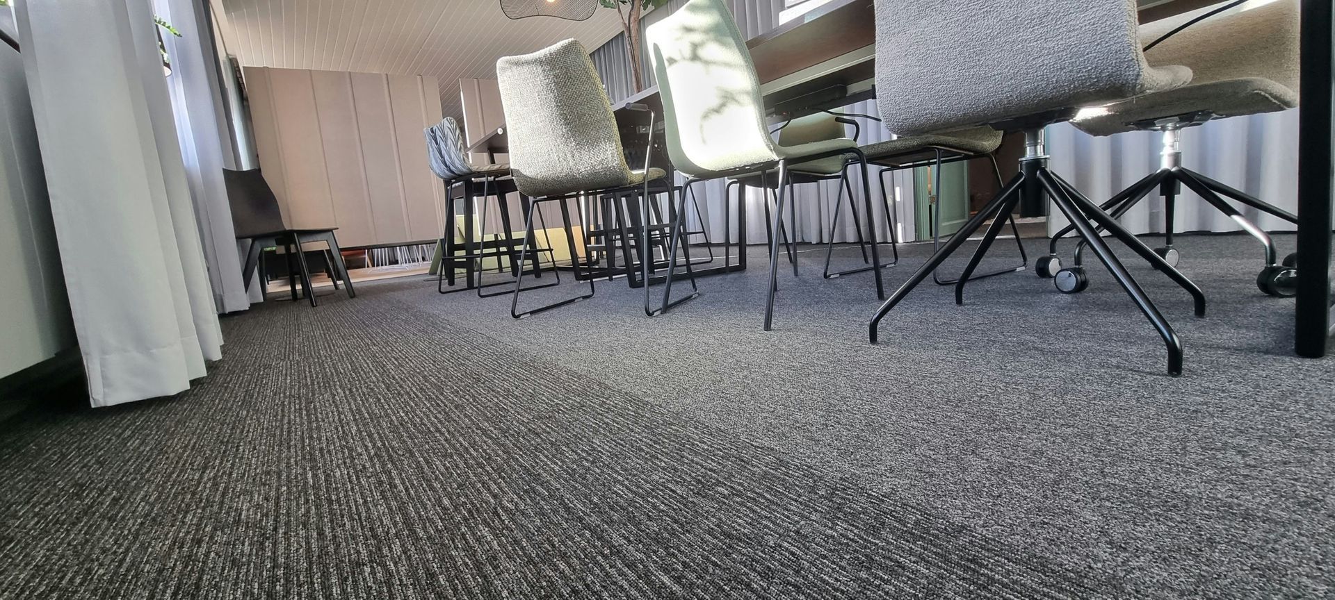 Low-angle view of a room with chairs and a textured gray carpet. A window with curtains is on the left.