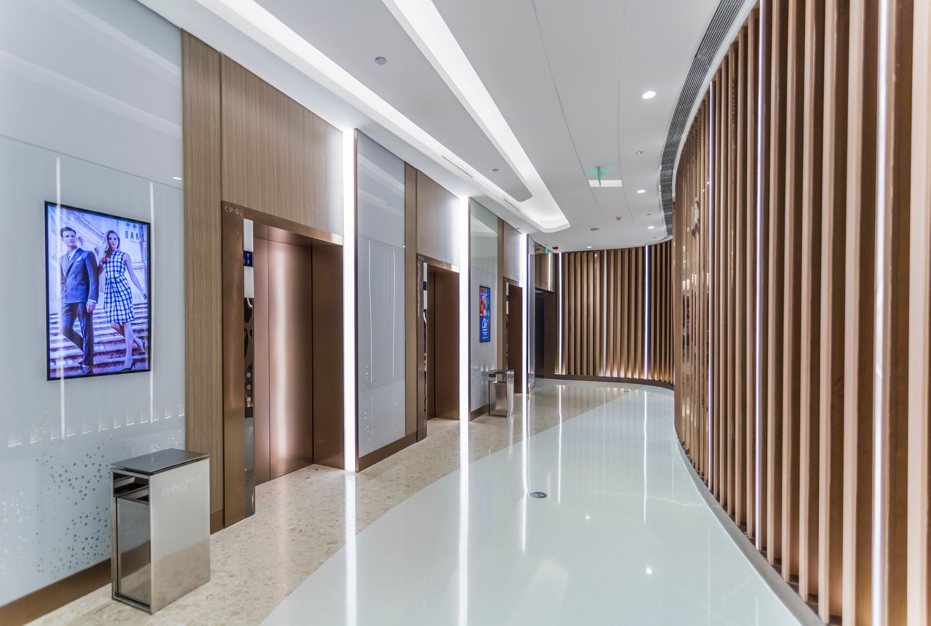 Elevator lobby with light wood paneling, three elevators, and a curved wall. White floors and recessed lighting.