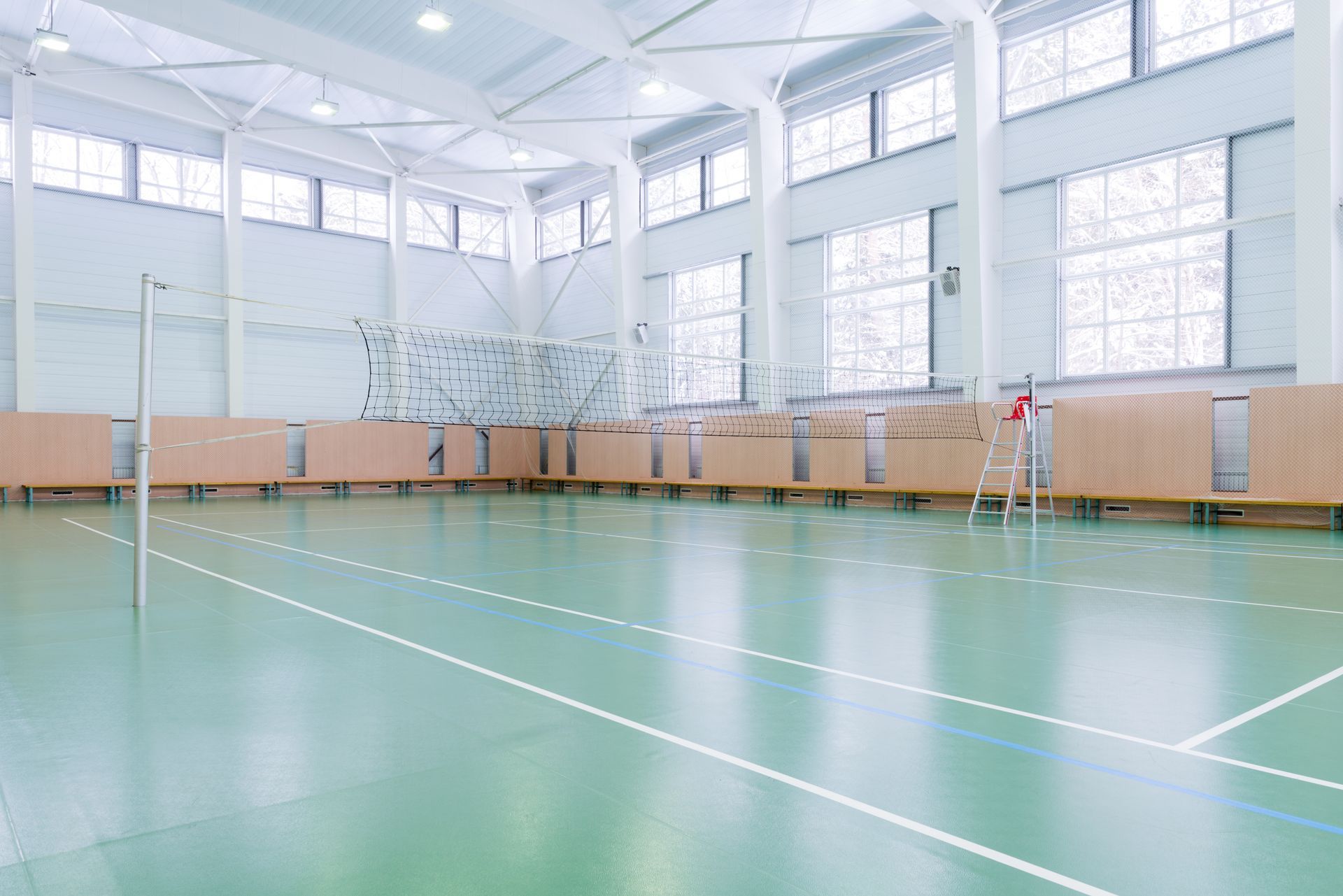 Empty indoor gymnasium with volleyball net. Light green floor, white lines, wooden side panels, large windows.