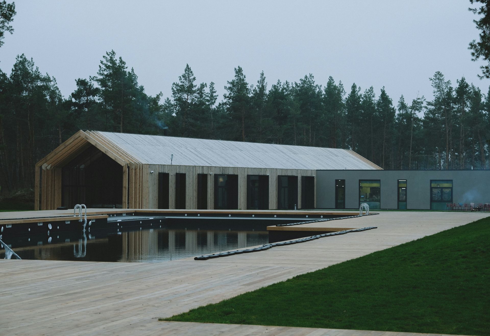 Modern wooden building next to a pool and lawn, with forest background.