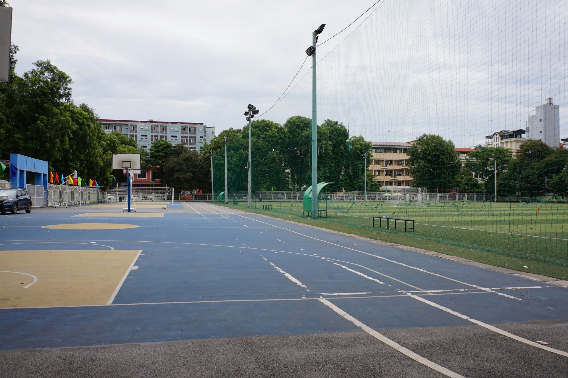 Blue sports court with a basketball hoop and a green field, trees, and buildings.