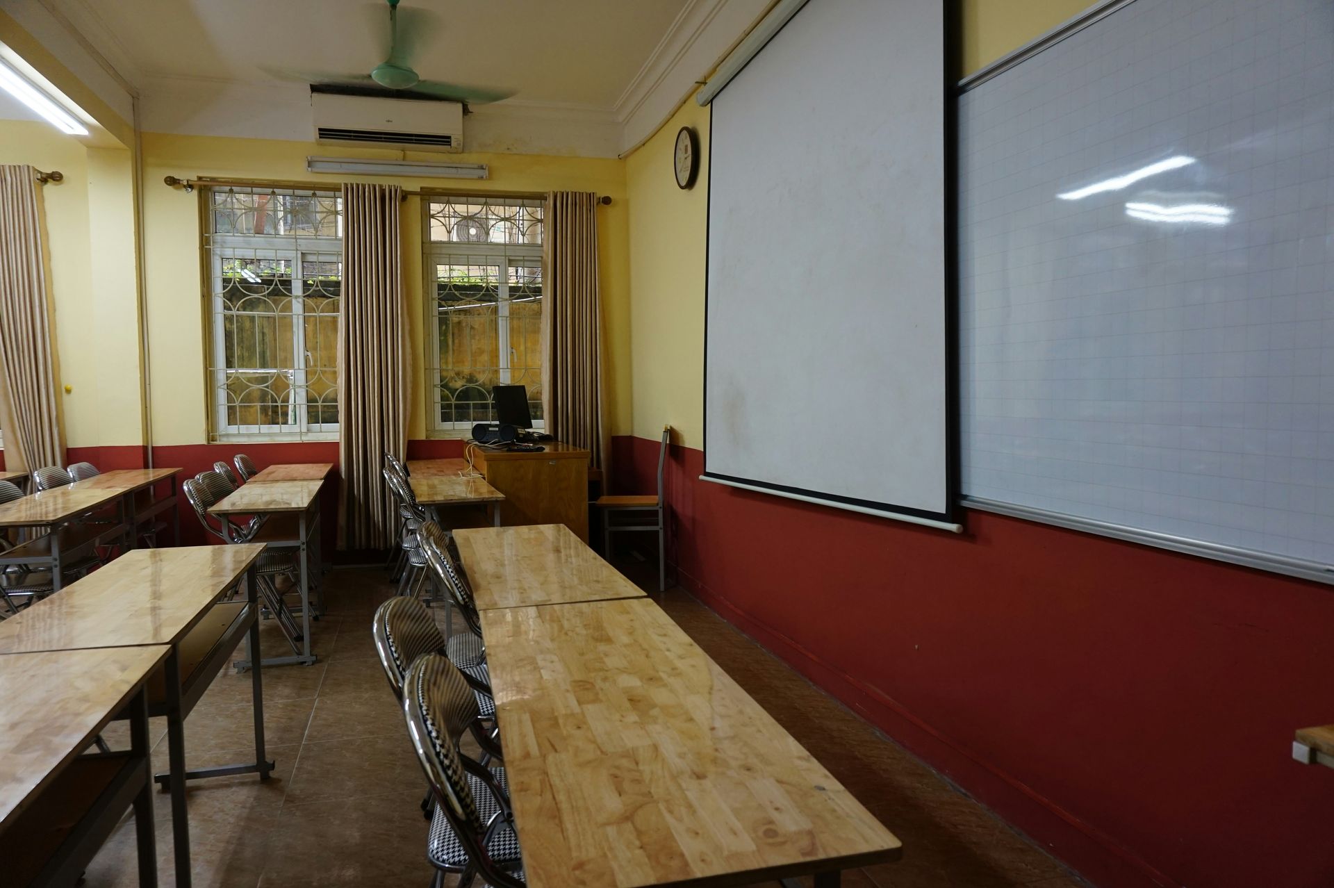 Classroom with desks, whiteboard, and projection screen. Windows with curtains and yellow walls.